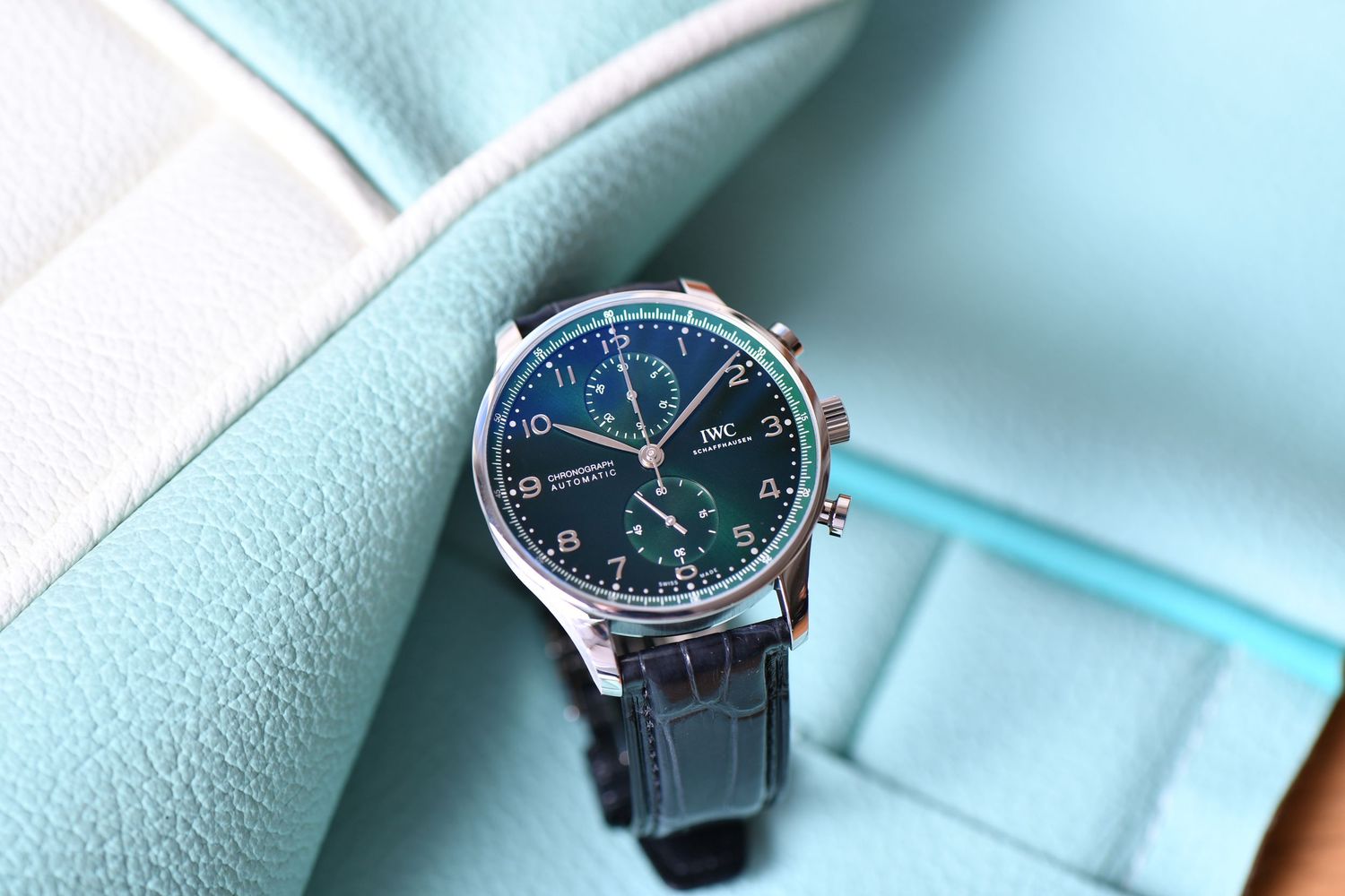 IWC Portugieser chronograph watch with green dial and blue leather strap on teal surface.