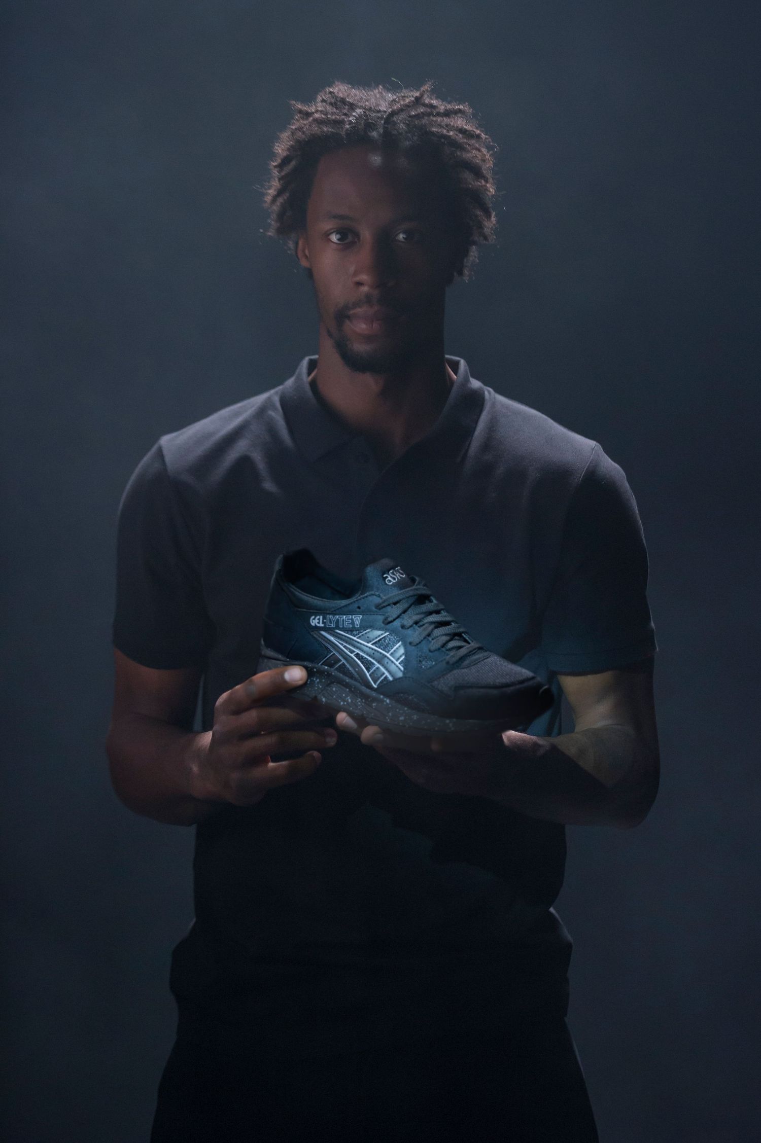 Gaël Monfils in black shirt holding glowing ASICS sneakers against dark background.