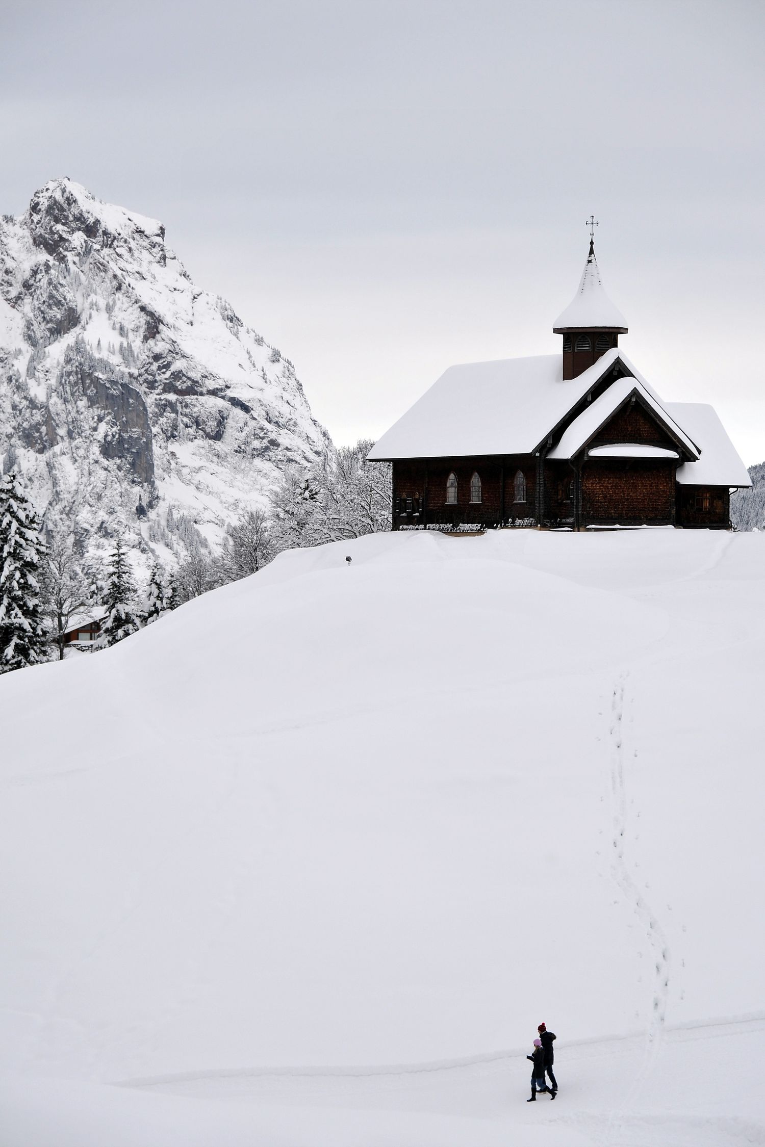 Snow-covered wooden chapel on a hillside with a lone figure walking through deep snow.