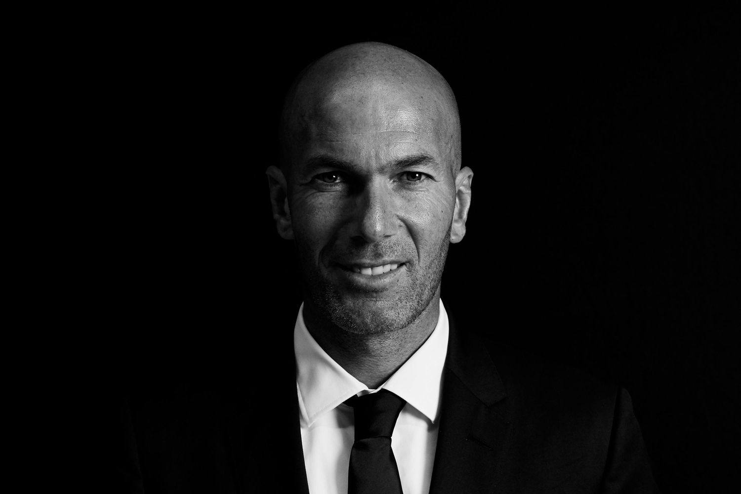 Zinedine Zidane in black suit and tie posing in dramatic black and white portrait photo.