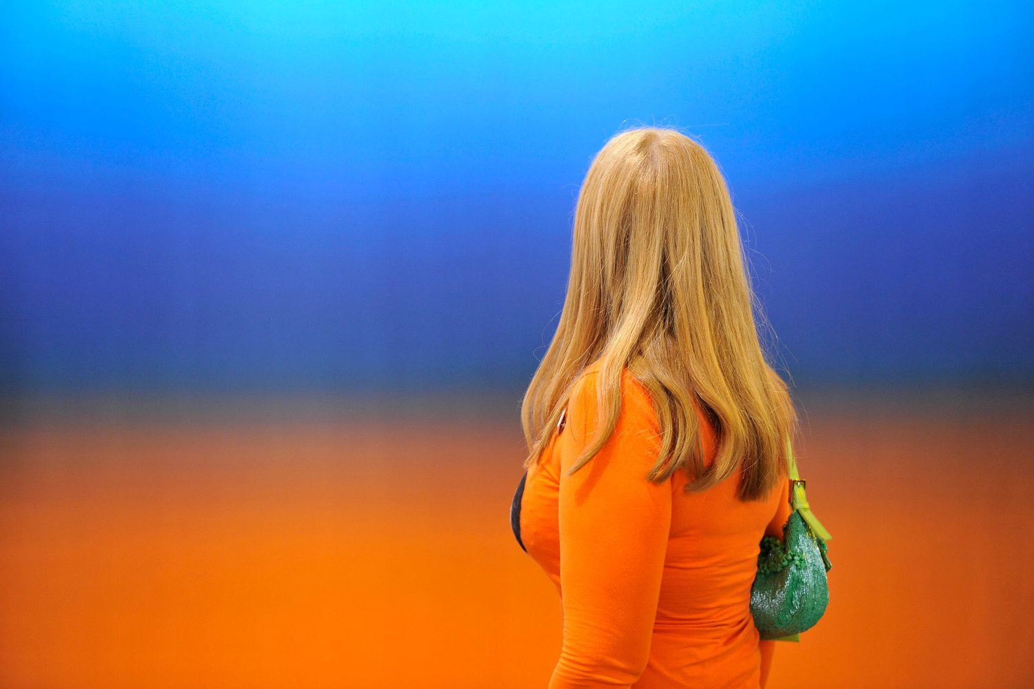Blonde woman in orange dress viewed from behind against colorful blue and orange background.