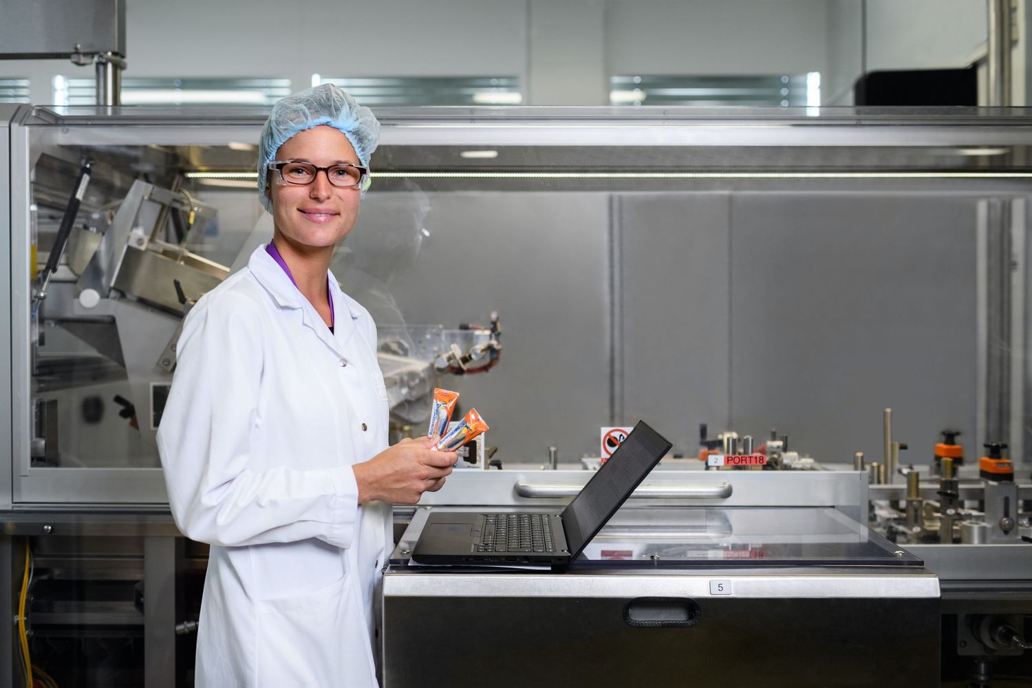 Female scientist in white lab coat holding laptop in modern pharmaceutical facility.