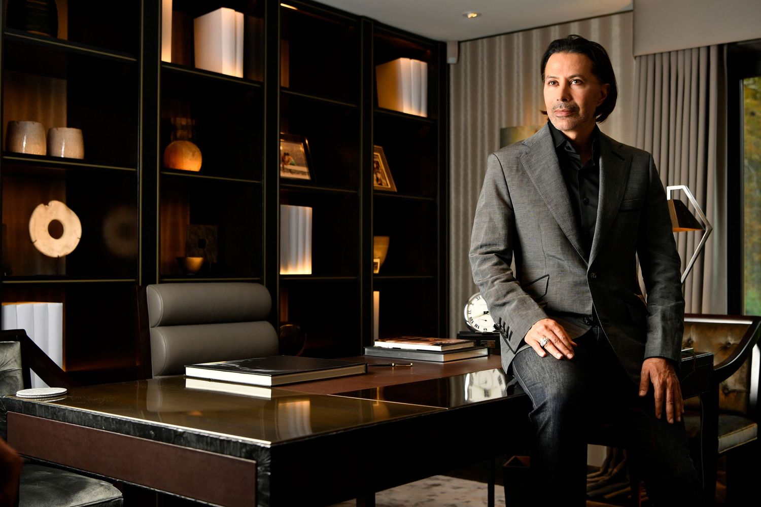 Confident man in grey blazer sitting on desk in elegant dark wood office setting.