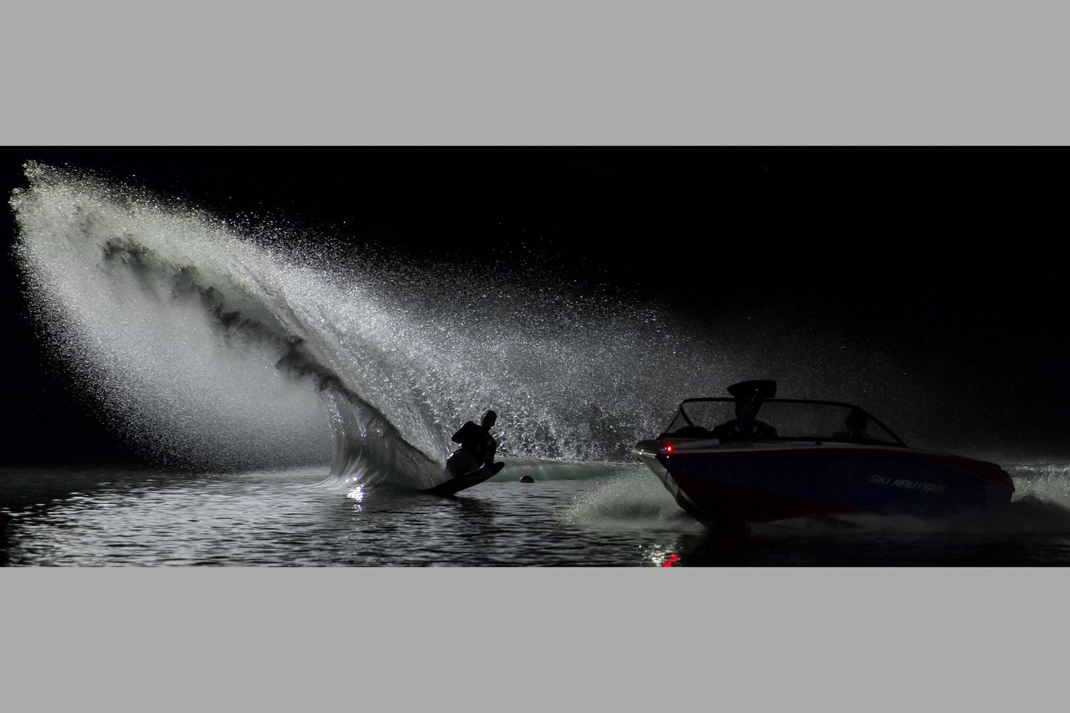 Night water skiing behind a speedboat creates dramatic water spray illuminated against a dark sky.