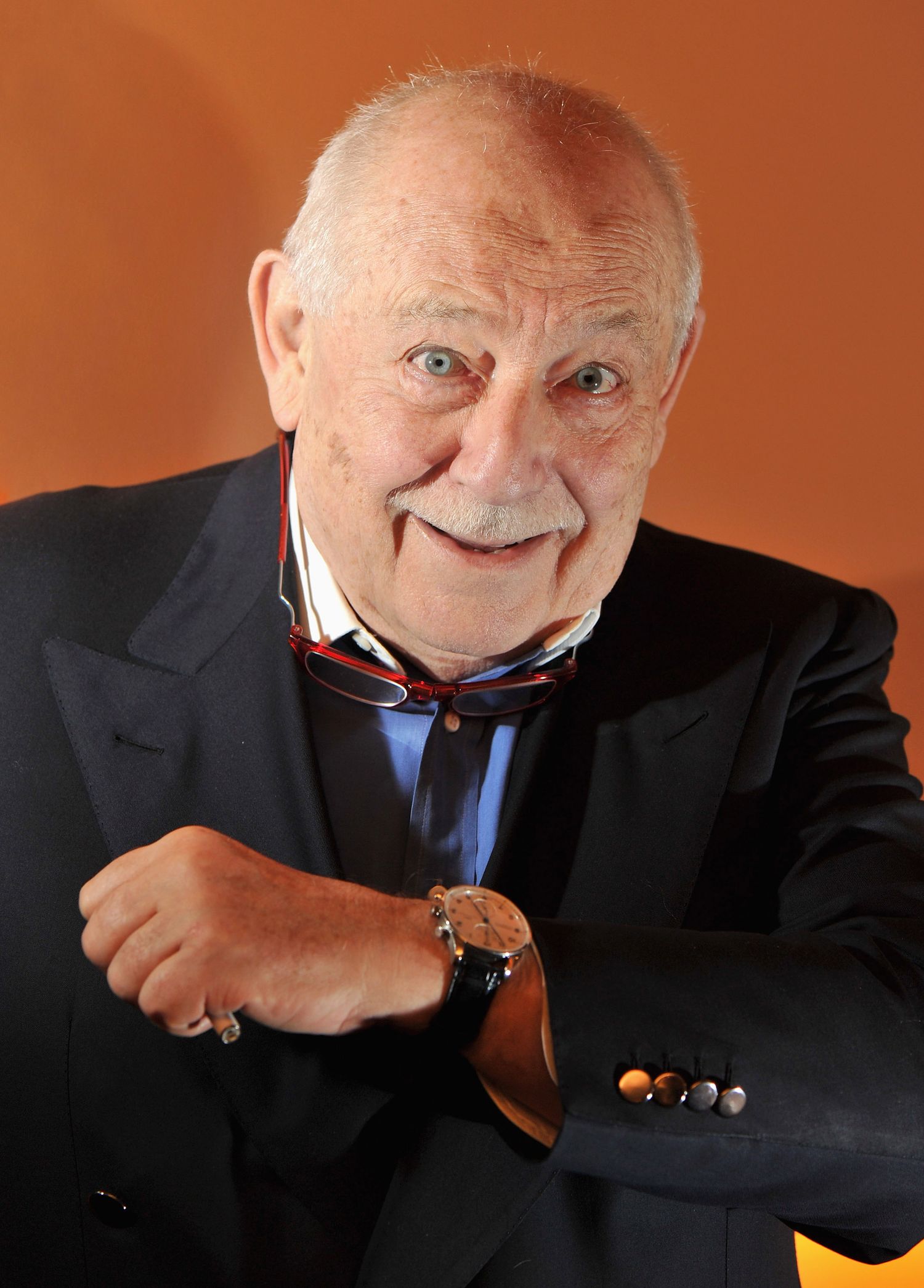 Franco Cologni in black suit displaying luxury watch against orange background.