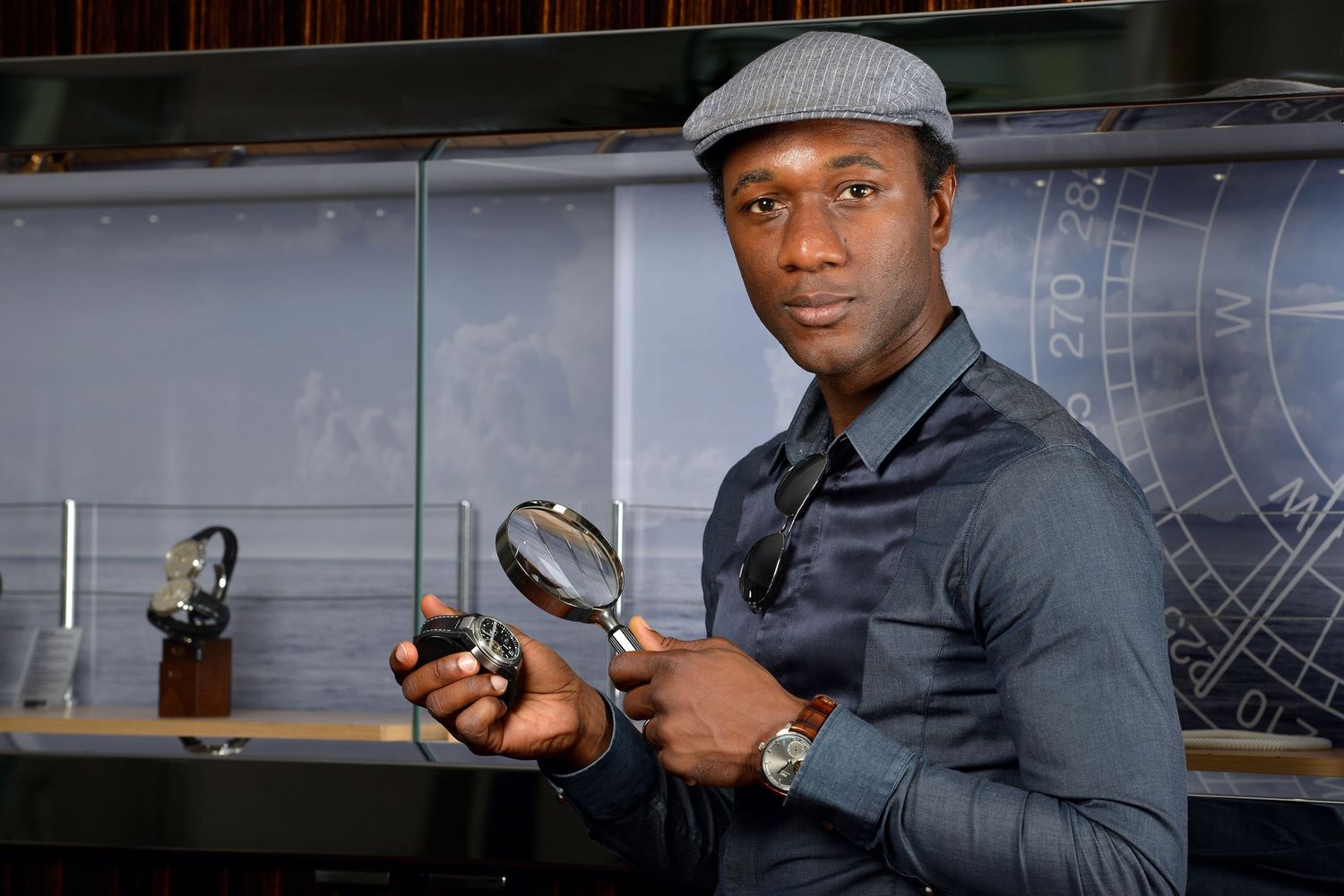 Aloe Blacc wearing a gray flat cap holds two magnifying glasses while posing indoors.