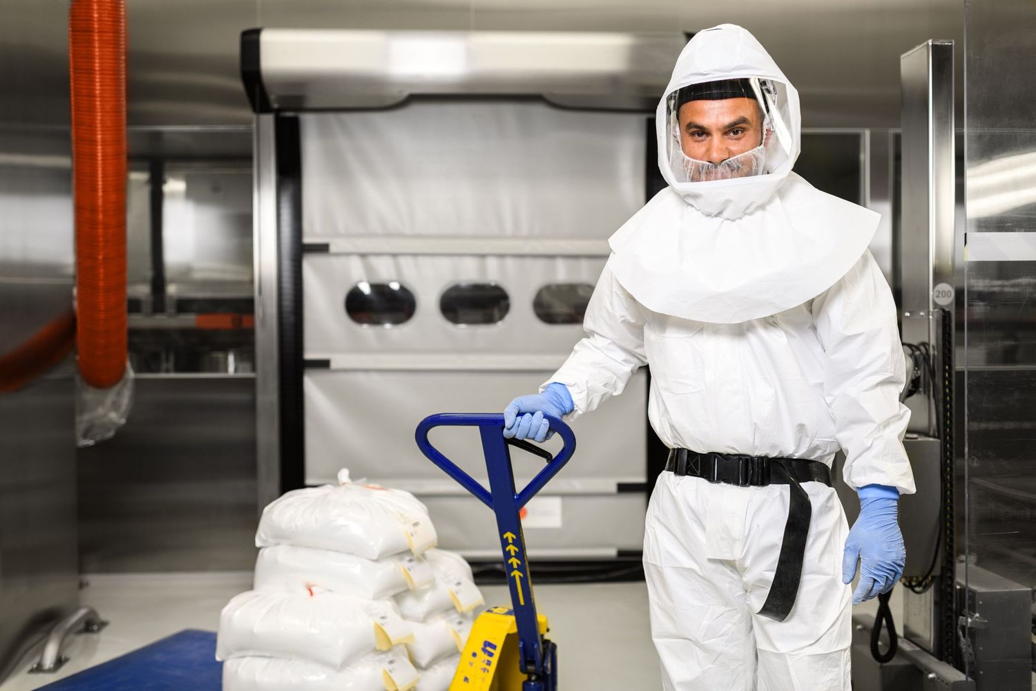 Worker in full white protective hazmat suit moving bags on a pallet jack.
