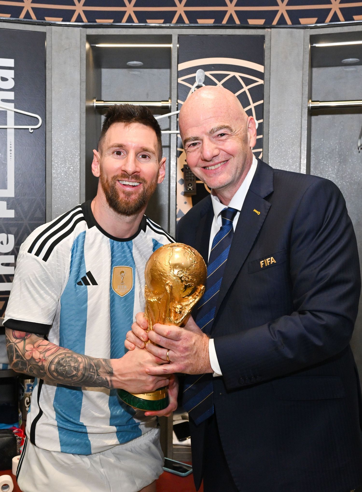 Two men proudly hold the FIFA World Cup trophy together in a locker room celebration after a championship win.