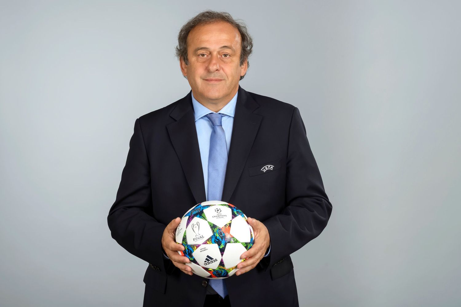 Michel Platini in a dark suit holds a UEFA Champions League soccer ball while posing for a photo.