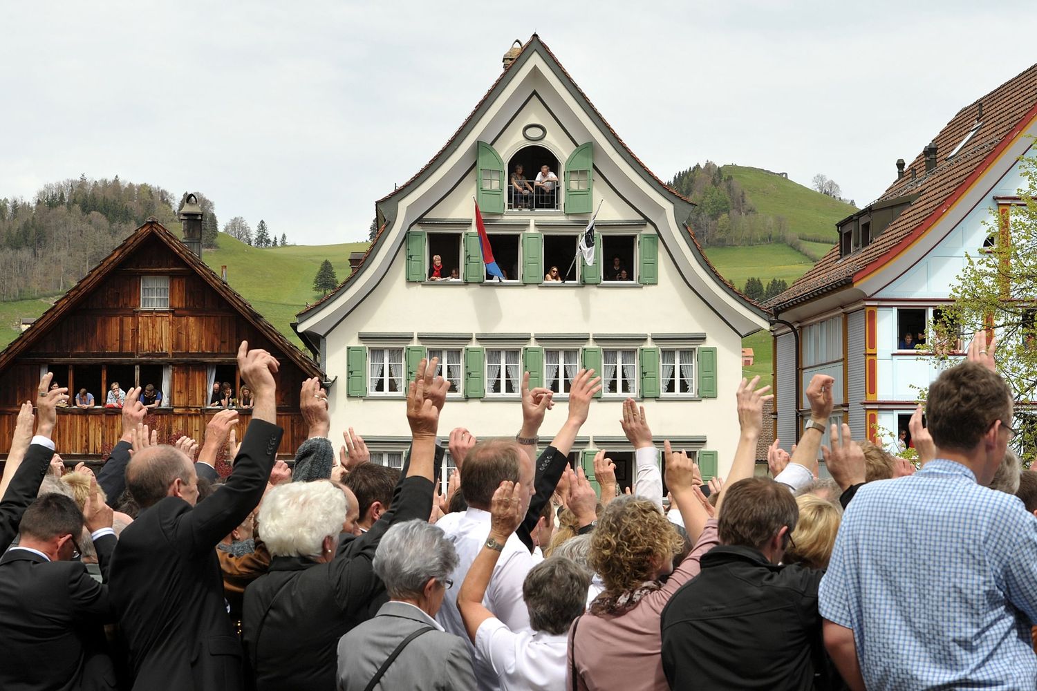 Crowd raising hands in Landsgemeinde vote outside a traditional Swiss chalet-style building during an outdoor assembly.