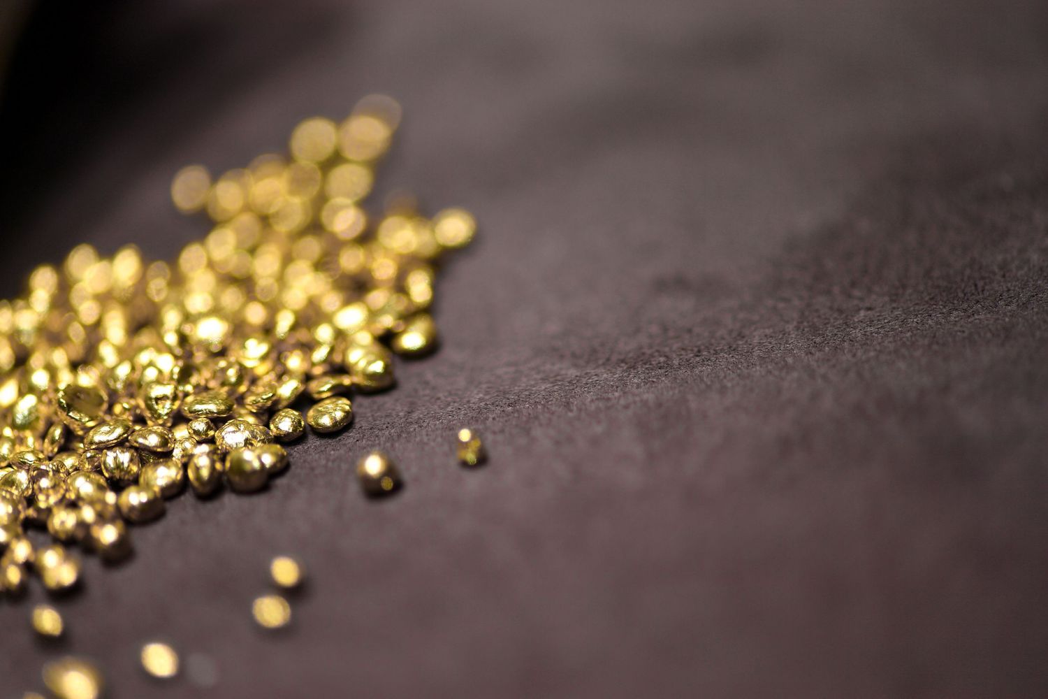 Scattered shiny gold granules on a dark textured surface with shallow depth of field.