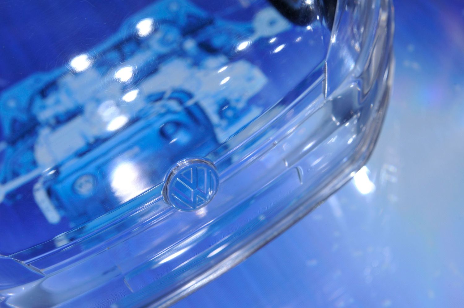 Close-up of a Volkswagen logo on a transparent car hood with blue lighting illuminating the engine.