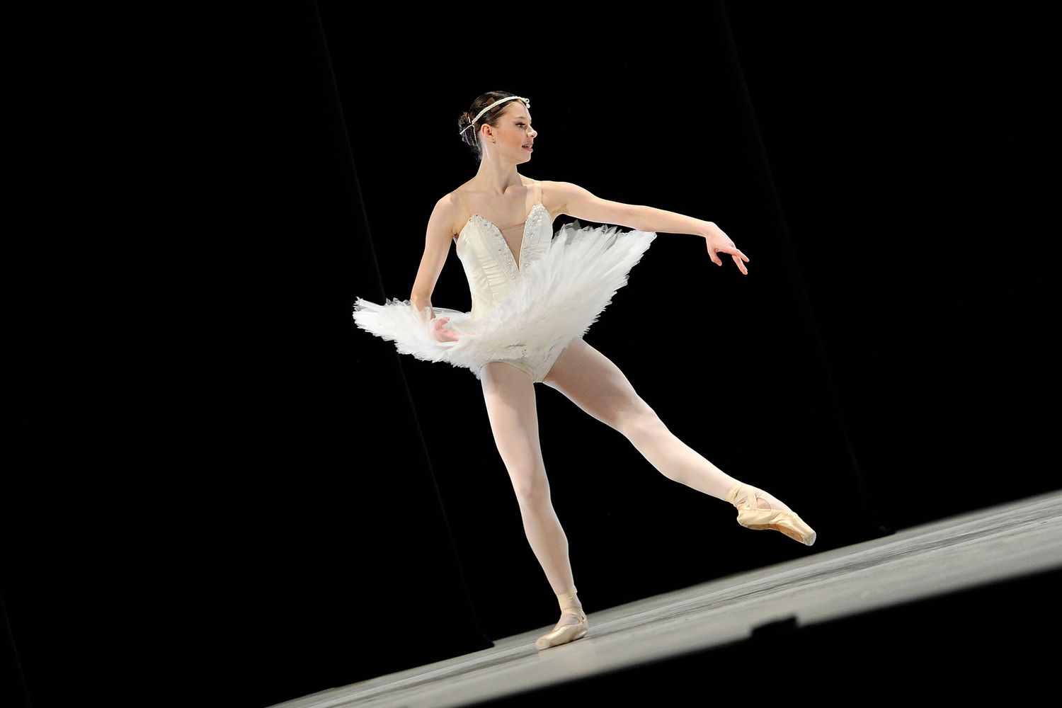Graceful ballerina performing in white tutu on stage against black background.