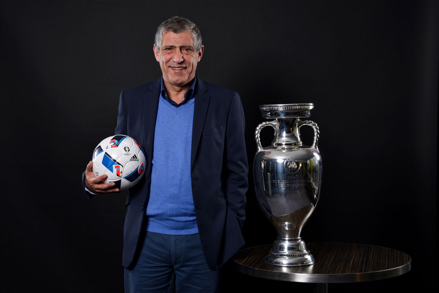 Man in blue blazer holding a soccer ball beside a large UEFA European Championship trophy on a dark background.