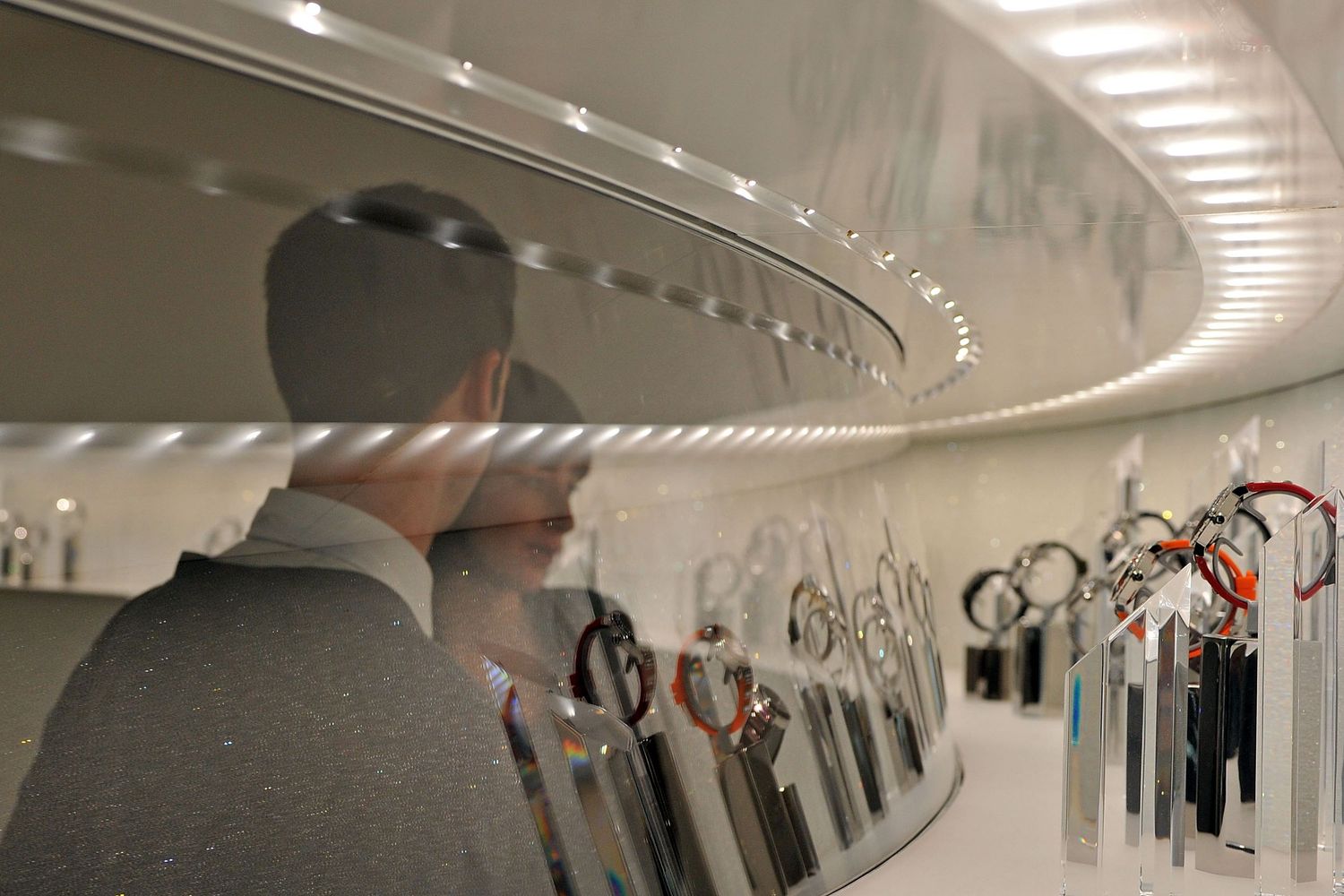 Man reflected in mirror surrounded by luxury watches displayed in a modern showroom.