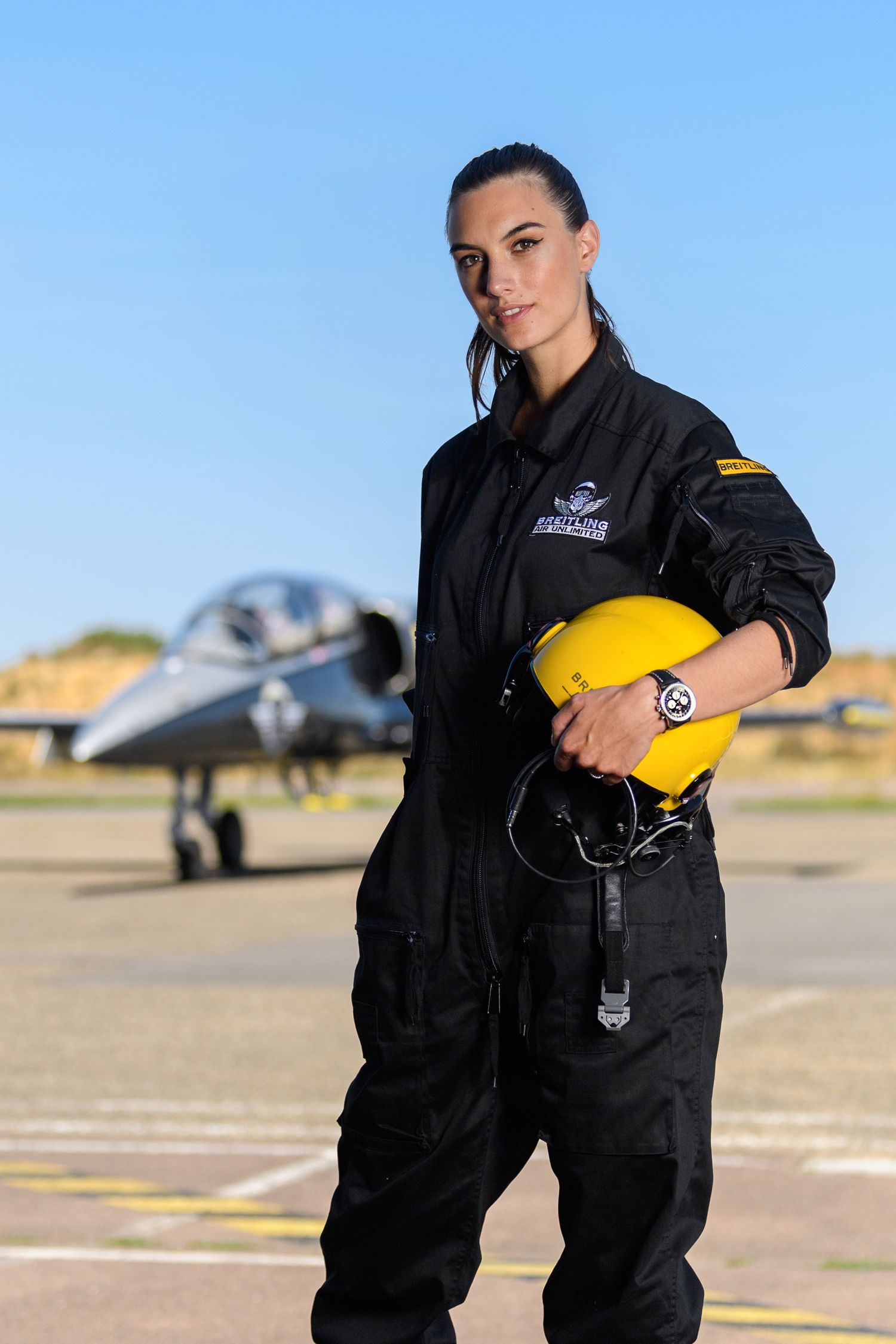 Female pilot in black flight suit holding yellow helmet on airport tarmac with jet behind her.