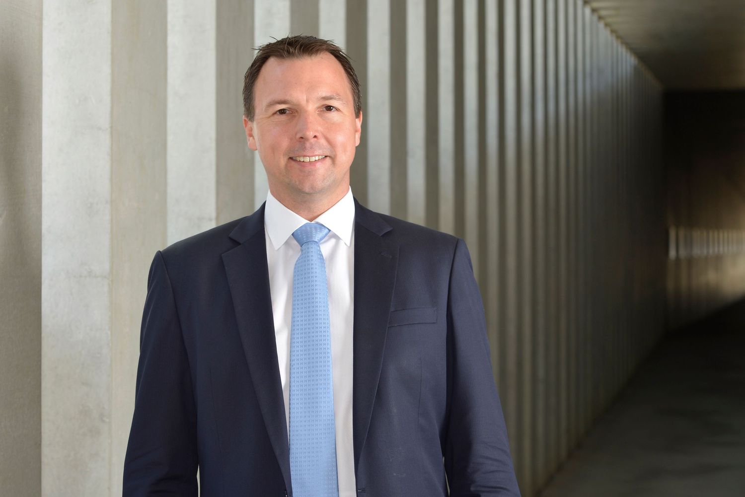 Businessman in navy suit with light blue tie posing in modern architectural corridor.