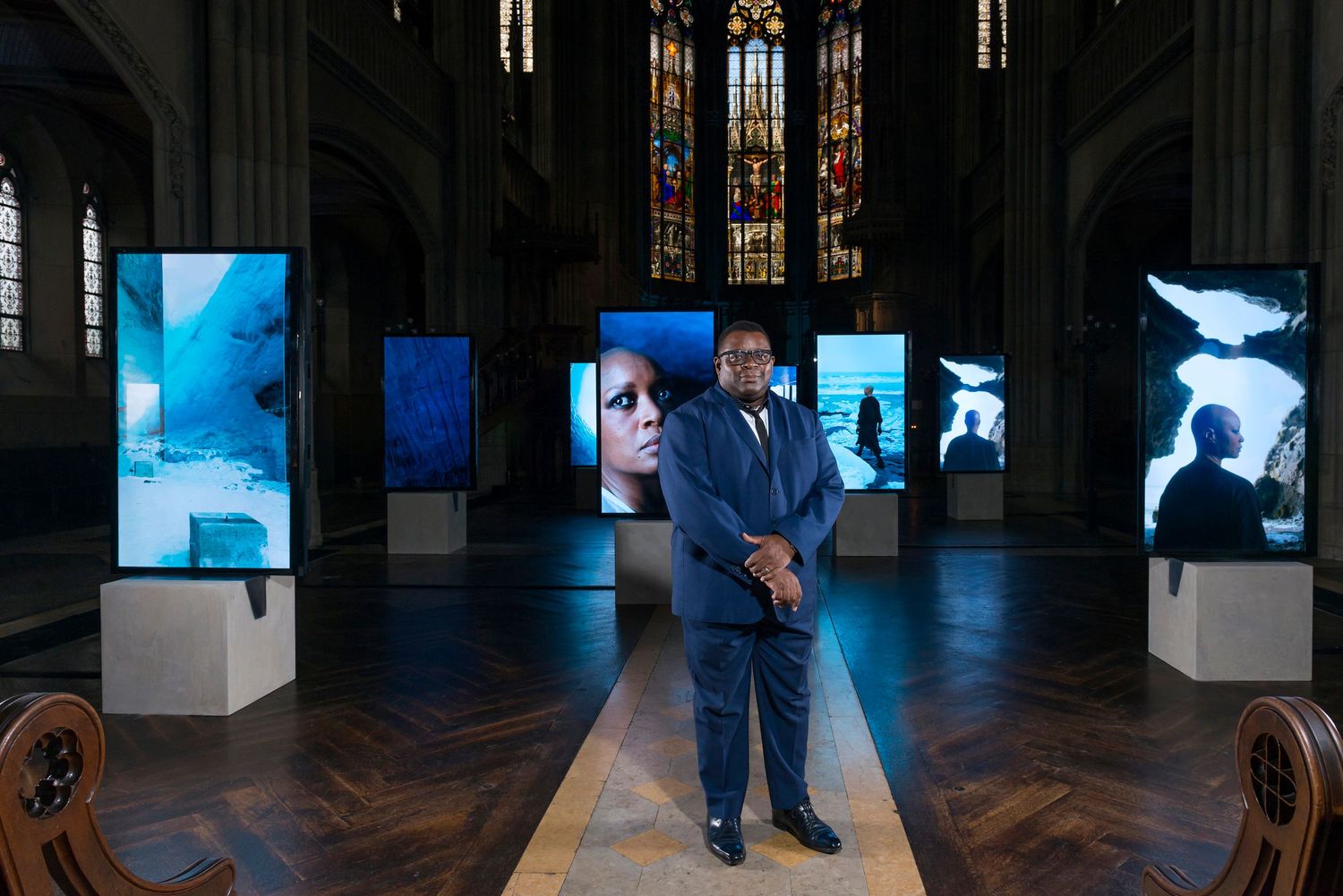 Artist Isaac Julien standing among illuminated display screens in a historic church with stained glass windows.