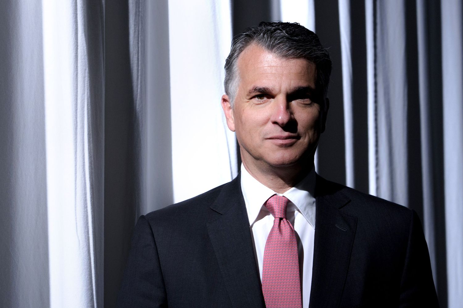 UBS CEO Sergio Ermotti posing for a portrait in dark suit and pink tie standing against striped background.