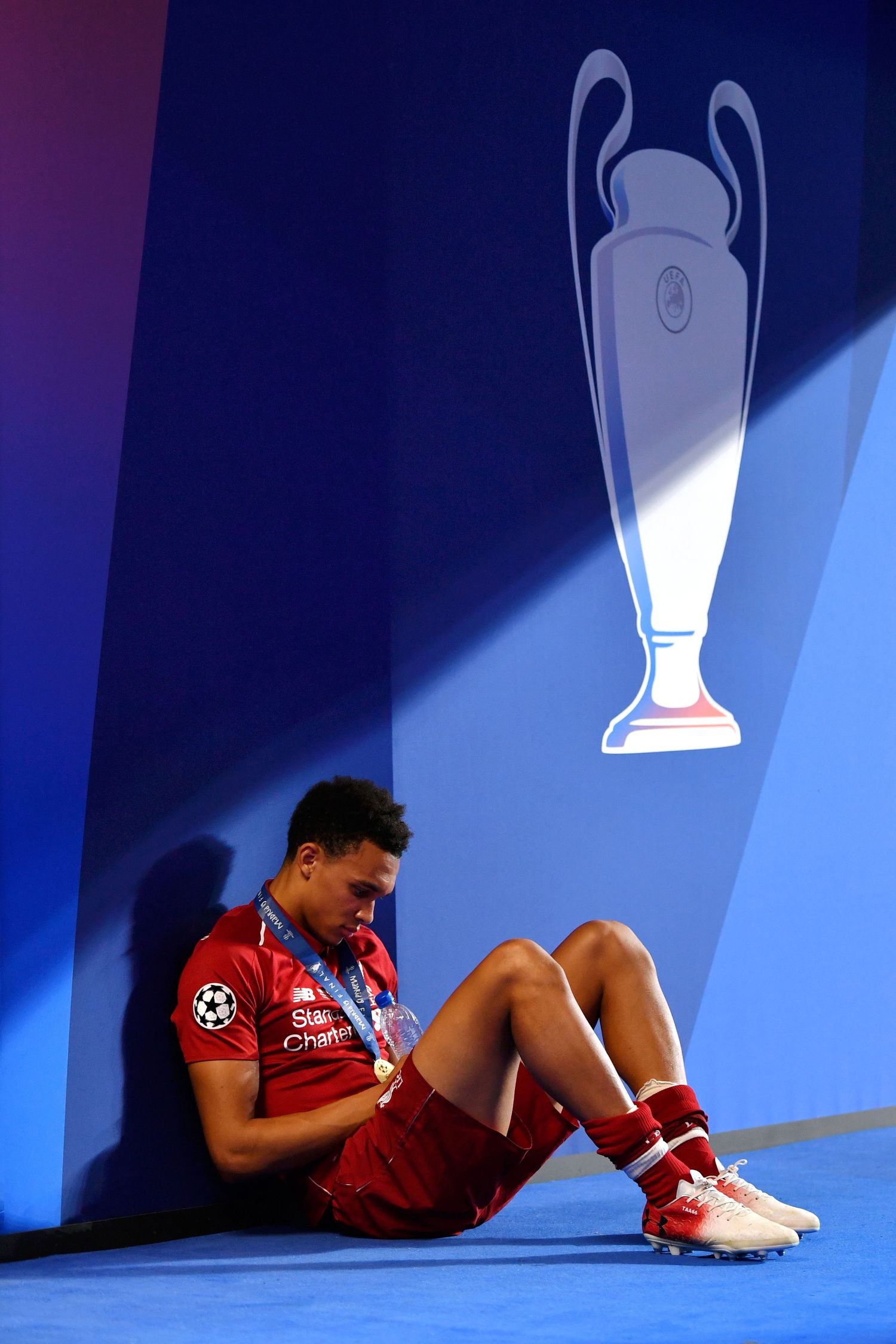 Liverpool footballer sits on floor wearing Champions League medal, leaning against blue wall with trophy graphic.
