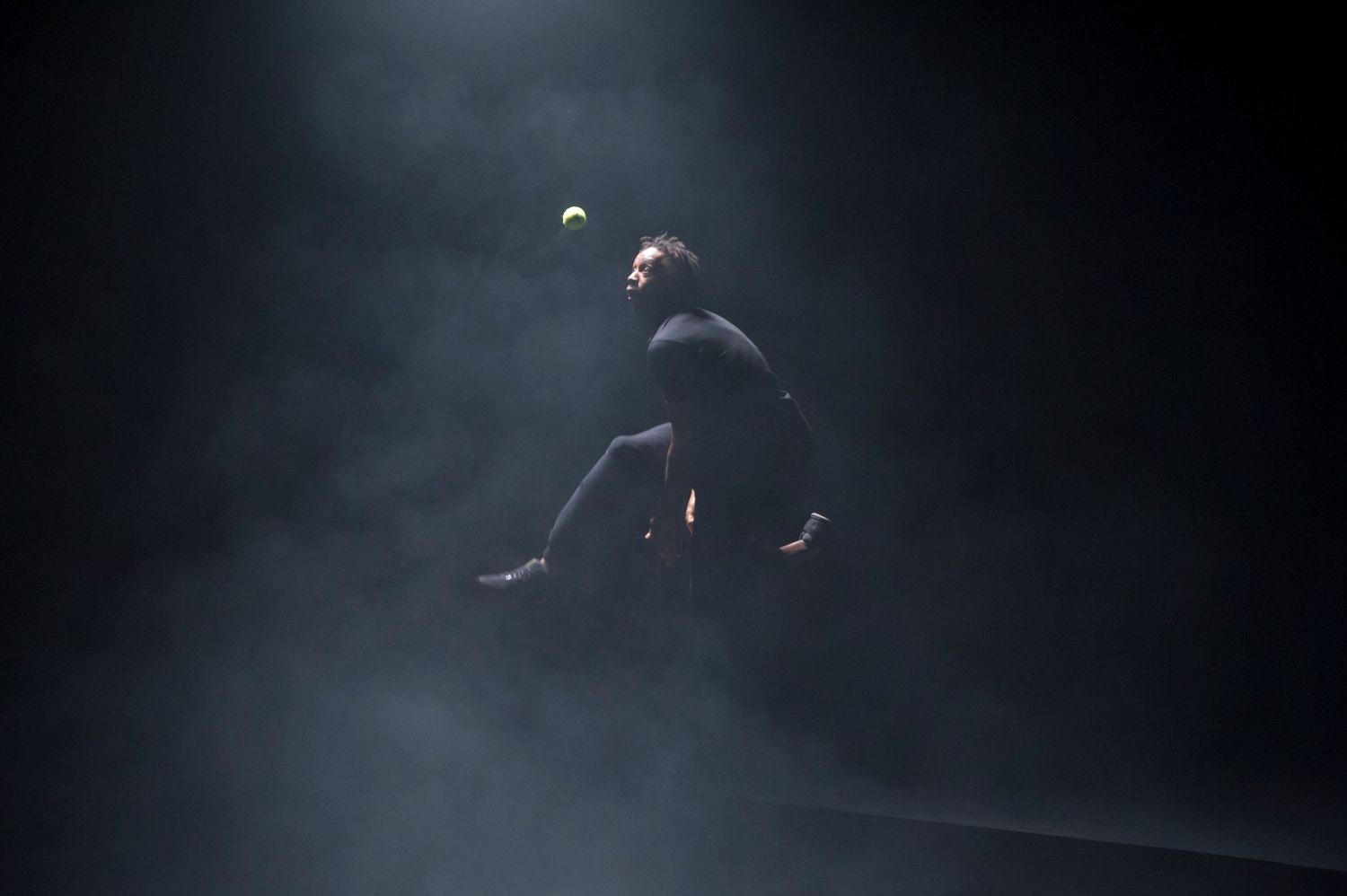 A tennis player leaps dramatically mid-air in a dark smoky arena, reaching for a ball illuminated by a spotlight.