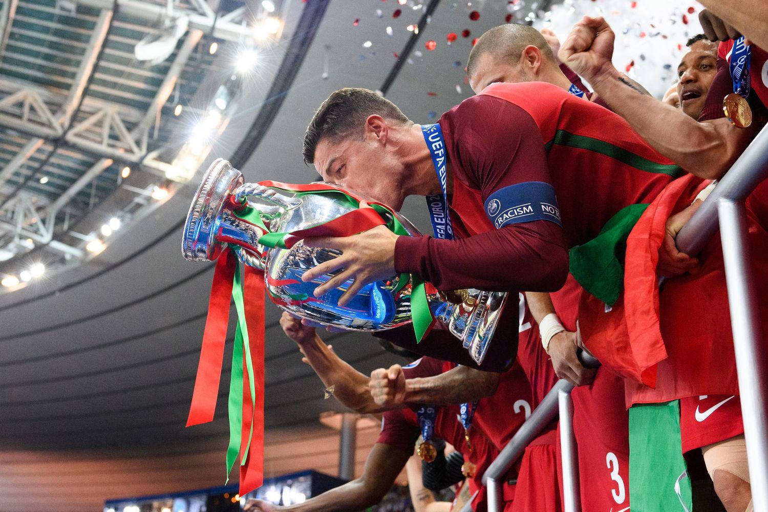 Portugal footballer kisses UEFA Euro Championship trophy while teammates celebrate in red jerseys.