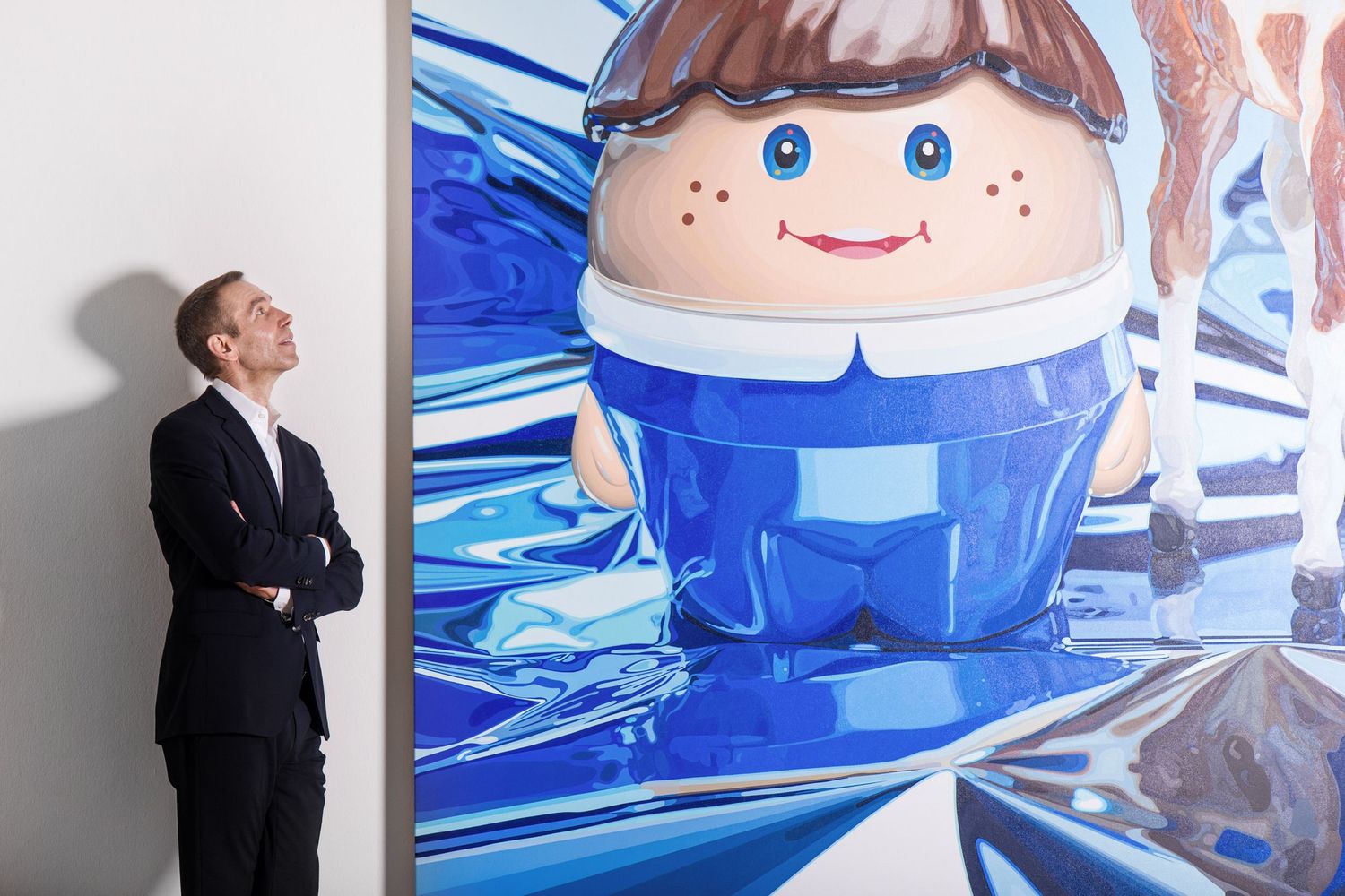 Jeff Koons in a dark suit stands with arms crossed beside a large colorful cartoon painting.