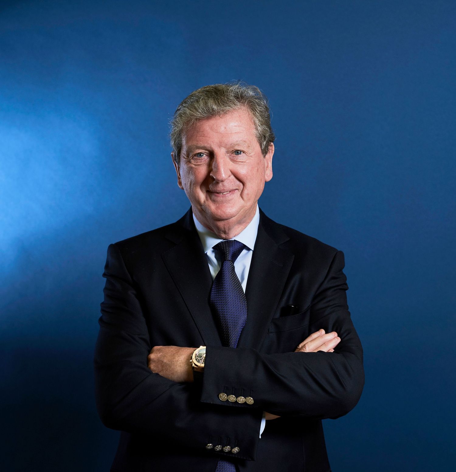 Roy Hodgson in a dark suit stands with arms crossed smiling against a blue background.