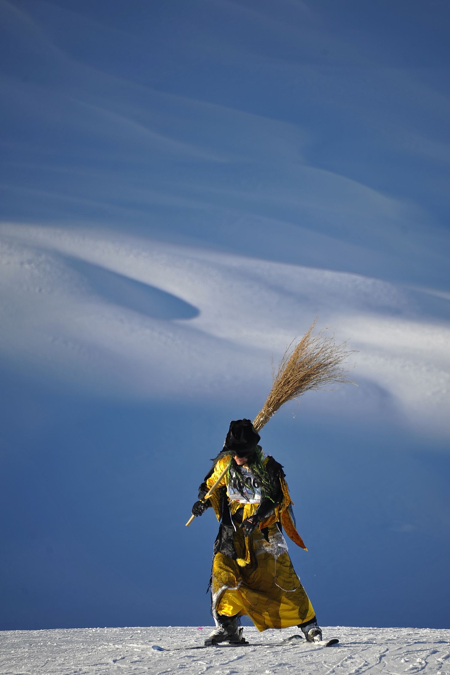 A performer in colorful traditional yellow costume carrying straw walks across a snowy alpine slope.