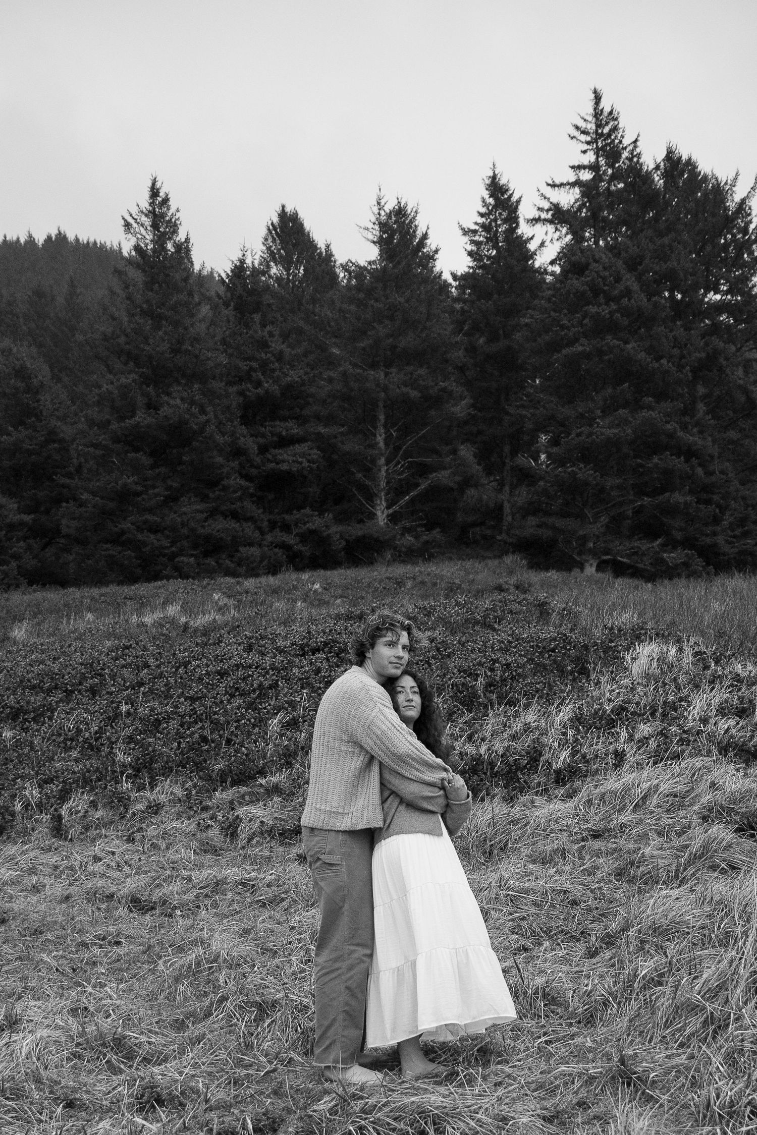 Black and white landscape photo of a couple walking through a misty field near evergreen trees.