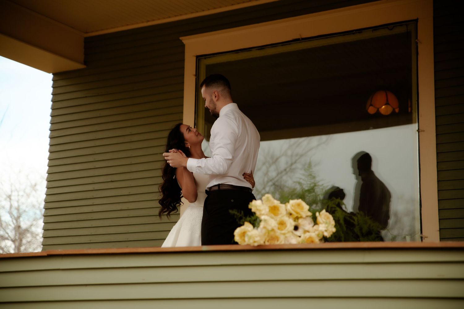 A bride and groom share a romantic embrace on a porch balcony with yellow flowers nearby.