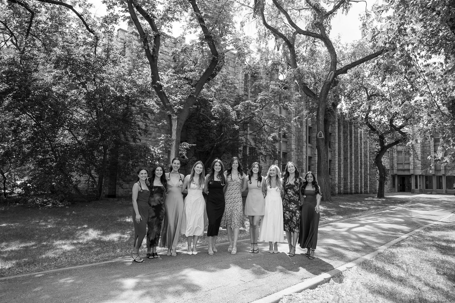 Black and white photo of a group of women posing together outdoors near a tree-lined path and building.