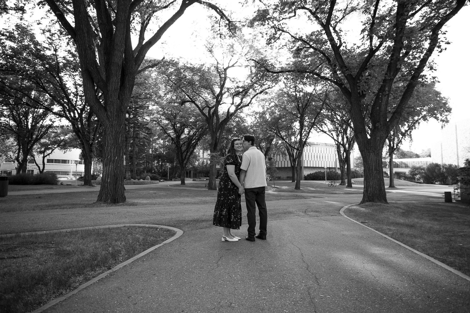 Black and white photo of a couple standing together on a curved path in a tree-lined park.