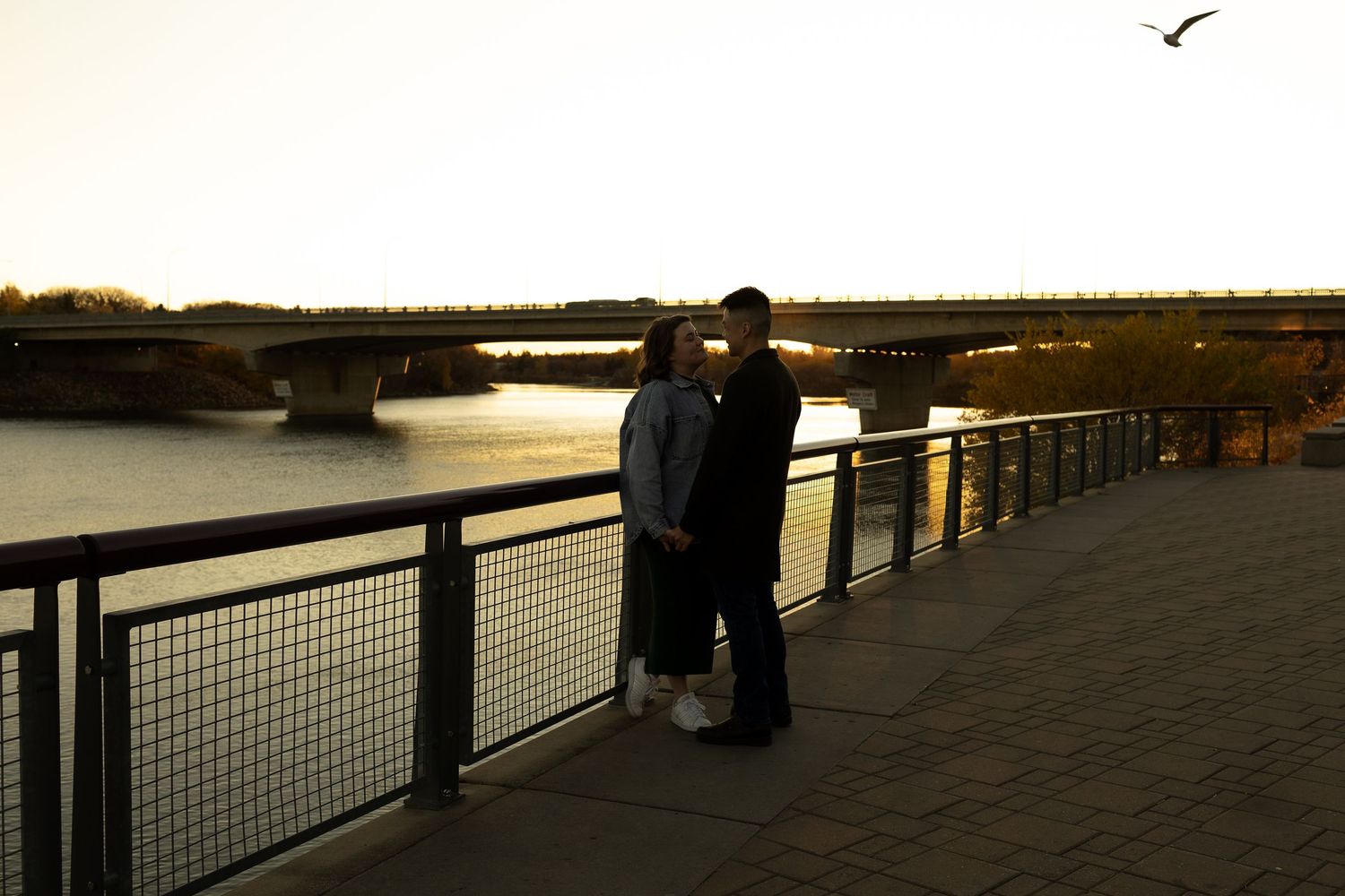 Couple sharing a romantic kiss on a waterfront bridge railing at golden sunset with river and bridge in background.