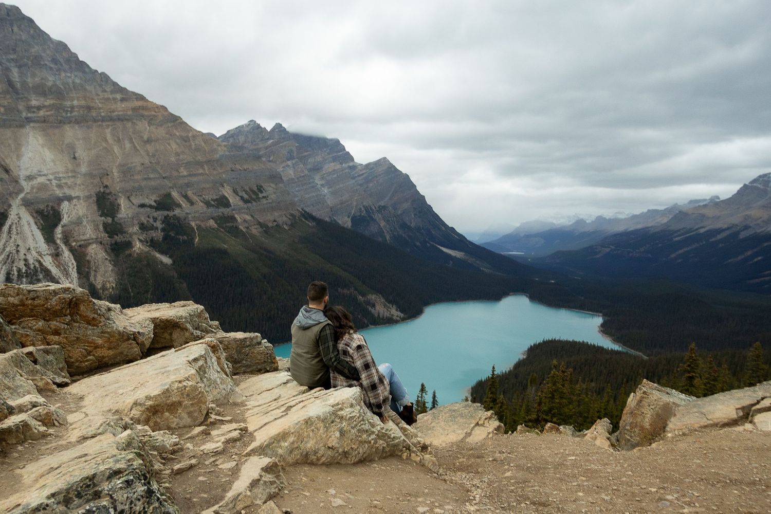 Two people sitting on rocky overlook admiring turquoise Peyto Lake surrounded by Canadian Rockies mountains.
