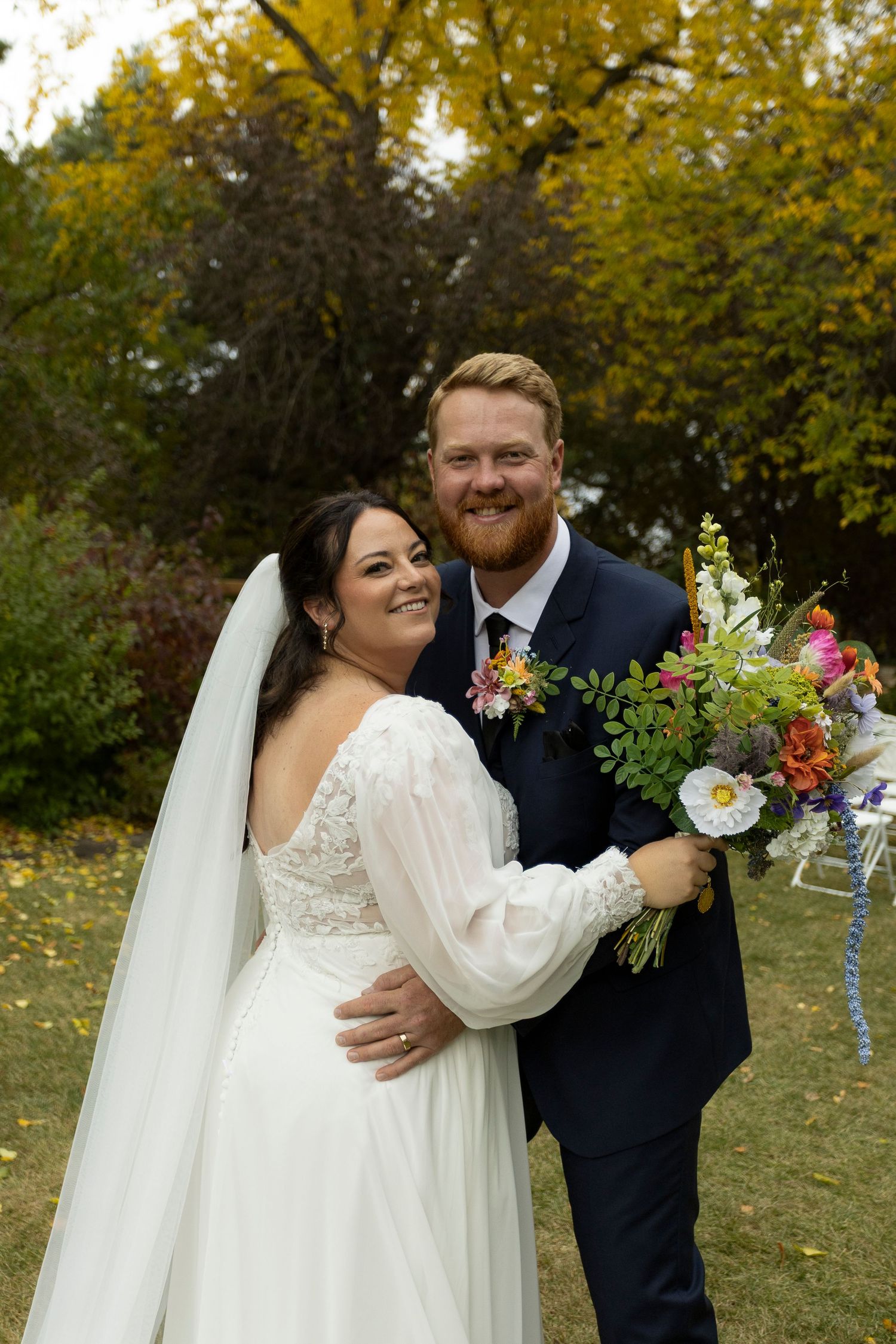Smiling bride in white lace gown with veil and groom in navy suit holding colorful bouquet outdoors in autumn setting.