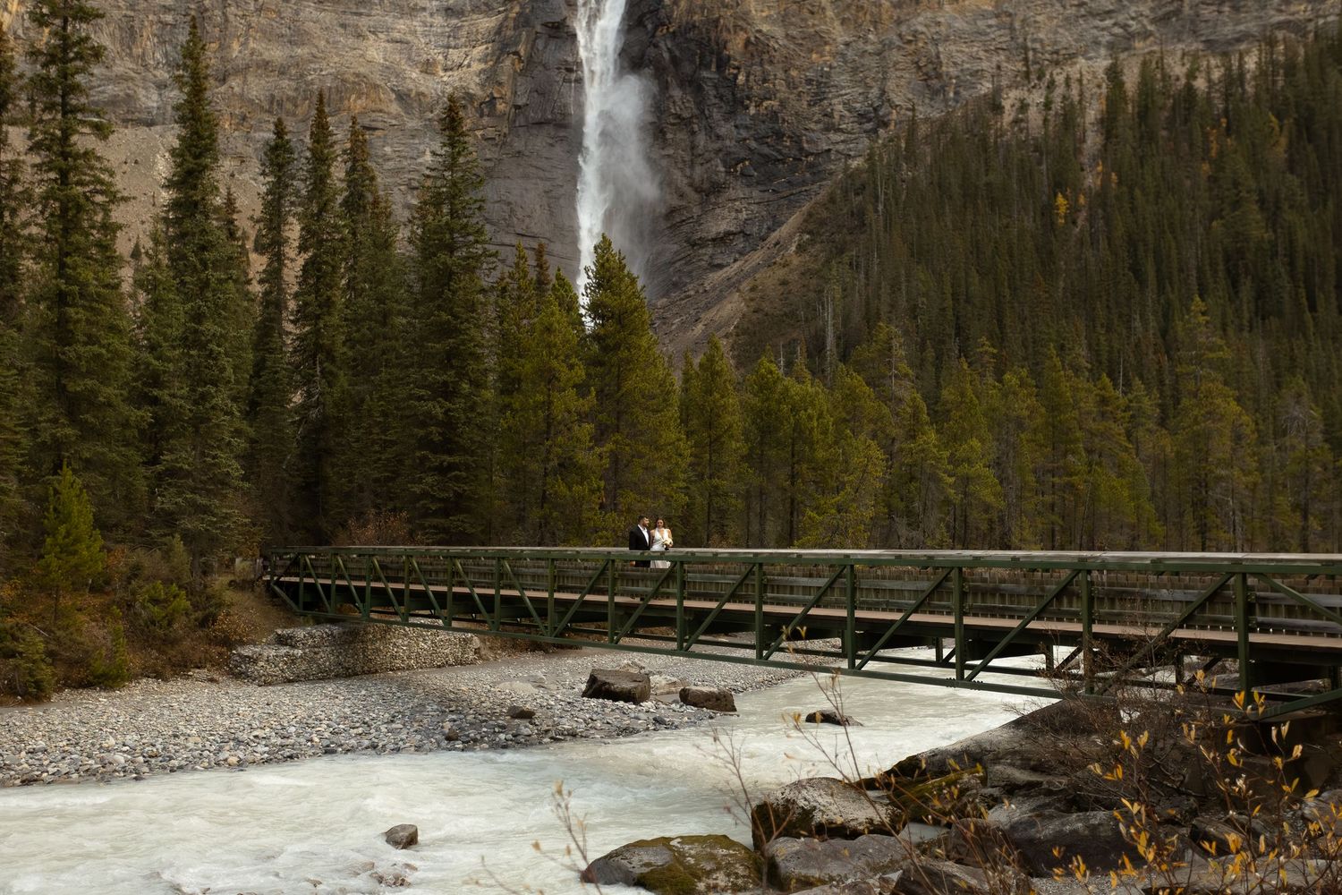Person on footbridge over glacial river with Takakkaw Falls and evergreen forest in Yoho National Park, Canada.