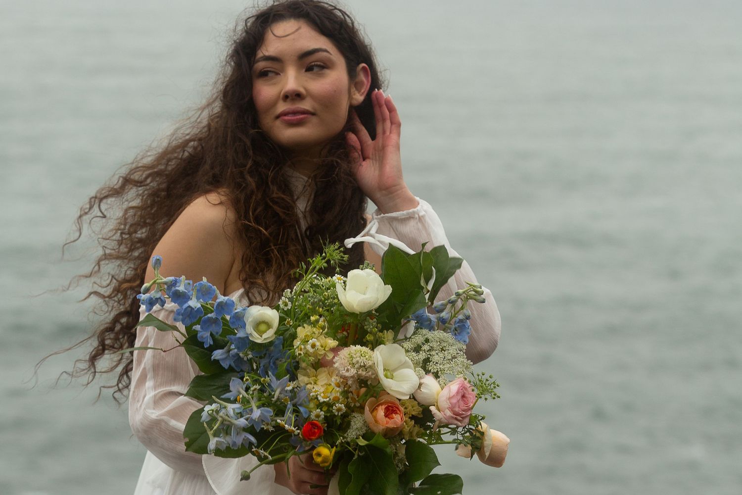 Woman with curly hair holding a colorful floral bouquet near the ocean in a white off-shoulder dress.