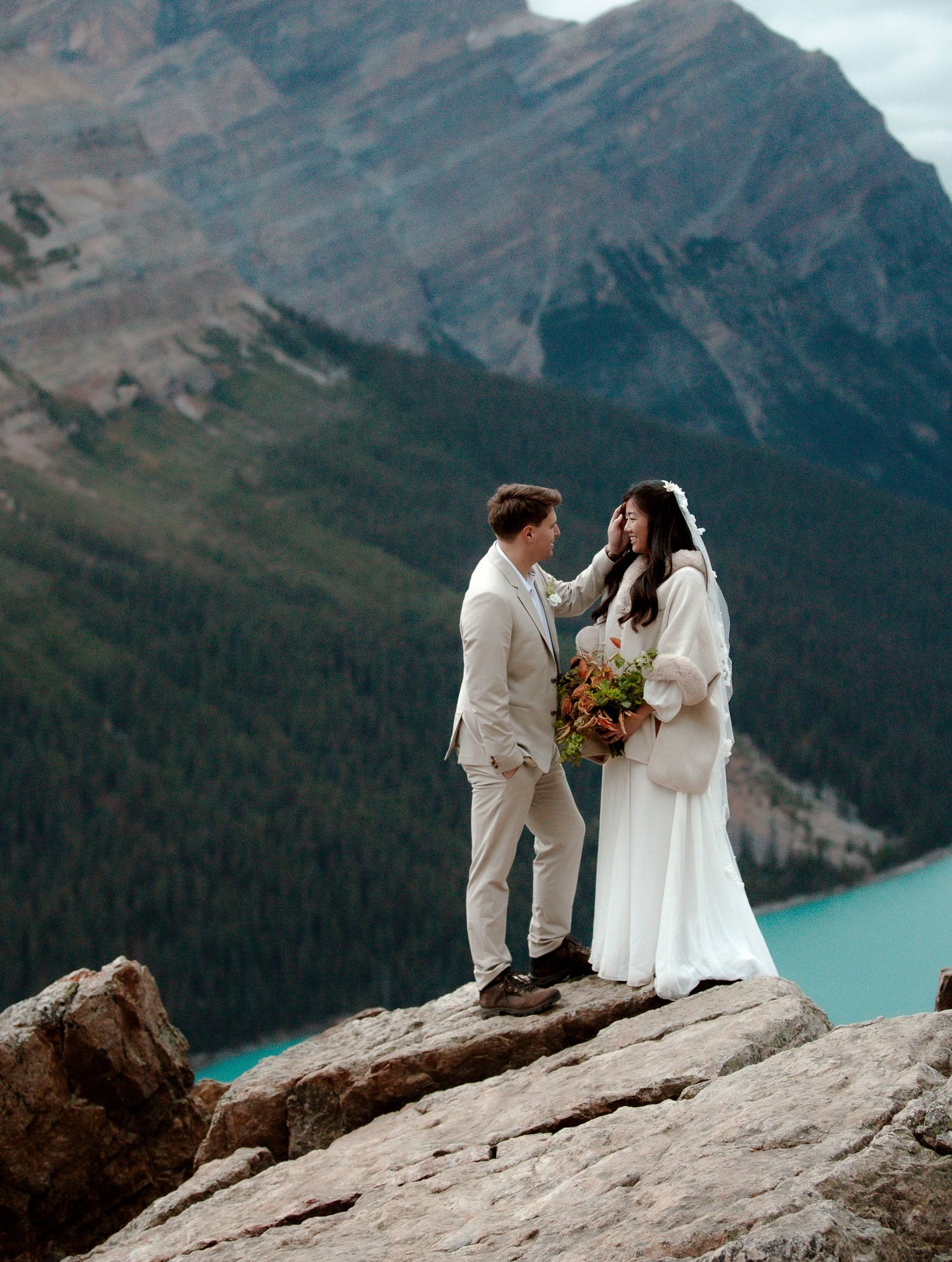 A couple shares an intimate moment on a rocky cliff overlooking turquoise Lake Louise in the Canadian Rockies.