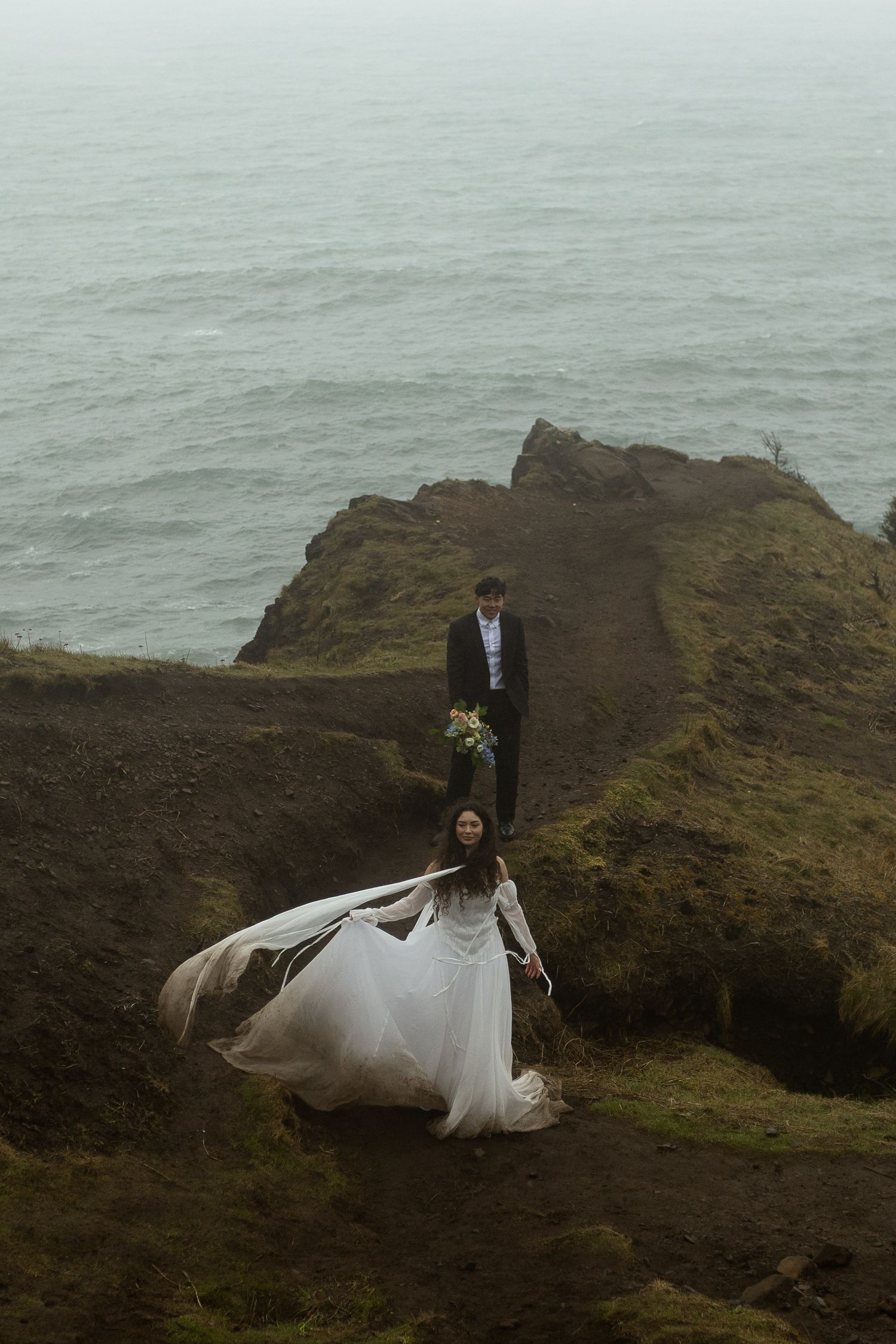 Bride in flowing white gown with veil blowing in wind on rocky coastal cliff, groom following behind on misty day.