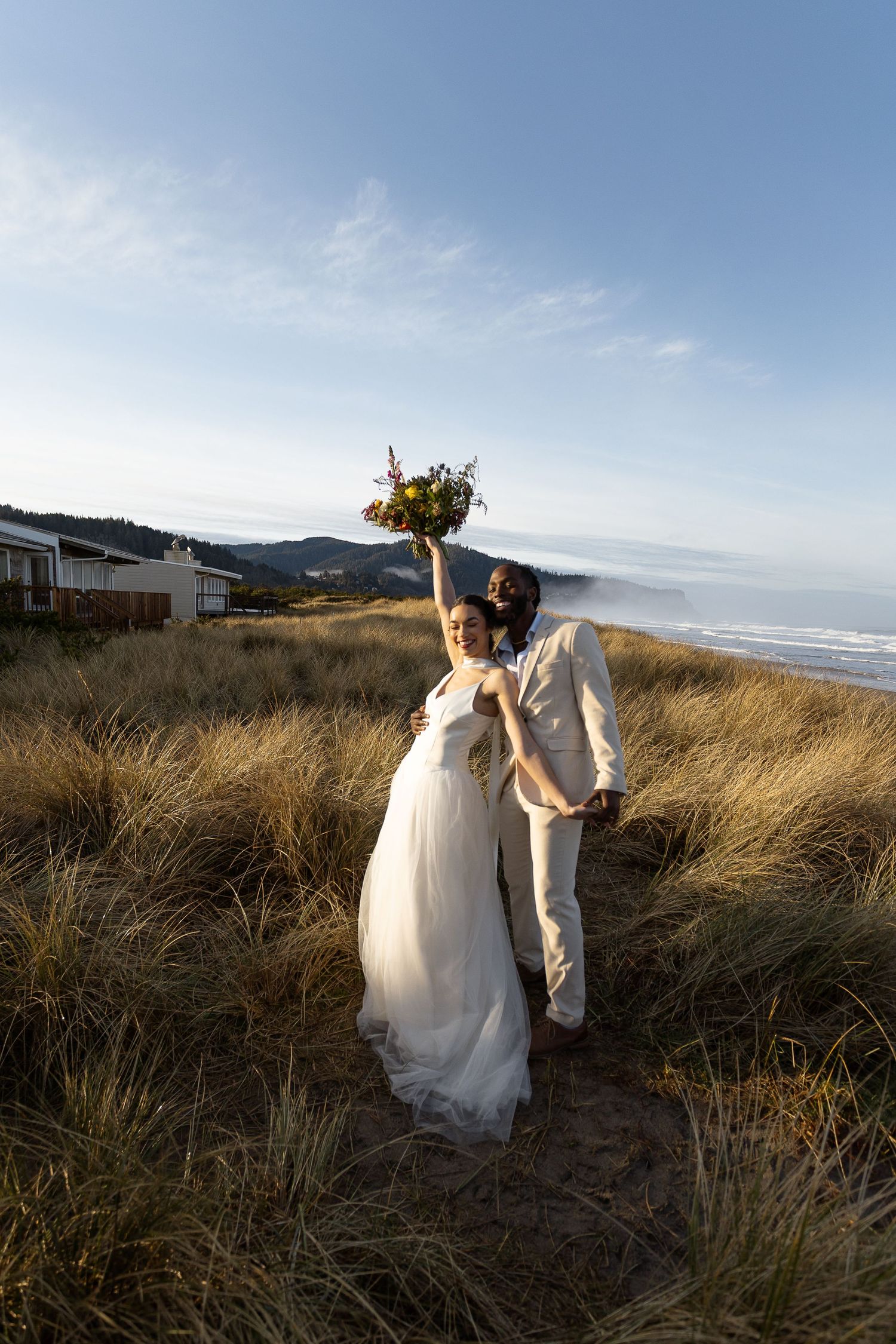 Bride raises bouquet on beach dunes as groom embraces her, ocean and golden grass in background.