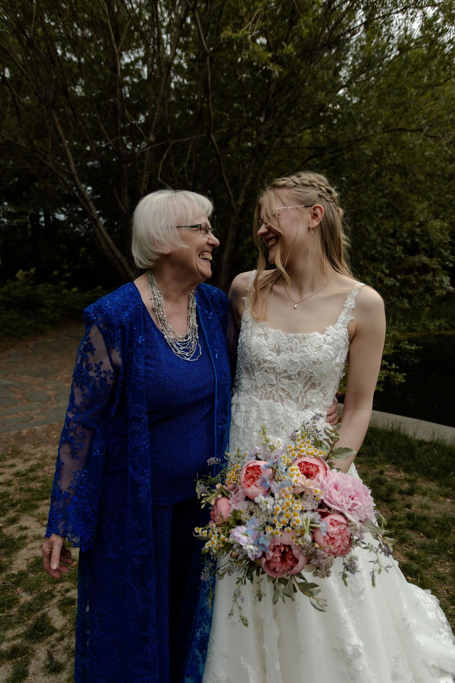A bride in a lace dress holds a pink bouquet while sharing a joyful moment outdoors at the University of Saskatchewan's boffin gardens.