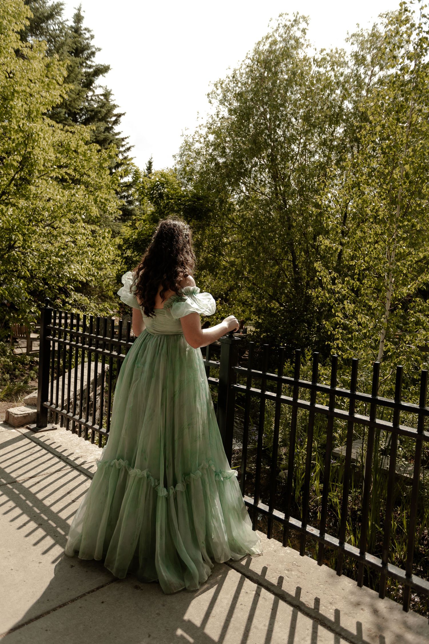 Woman in elegant green tulle gown standing on sunlit balcony overlooking lush trees and black iron fence.