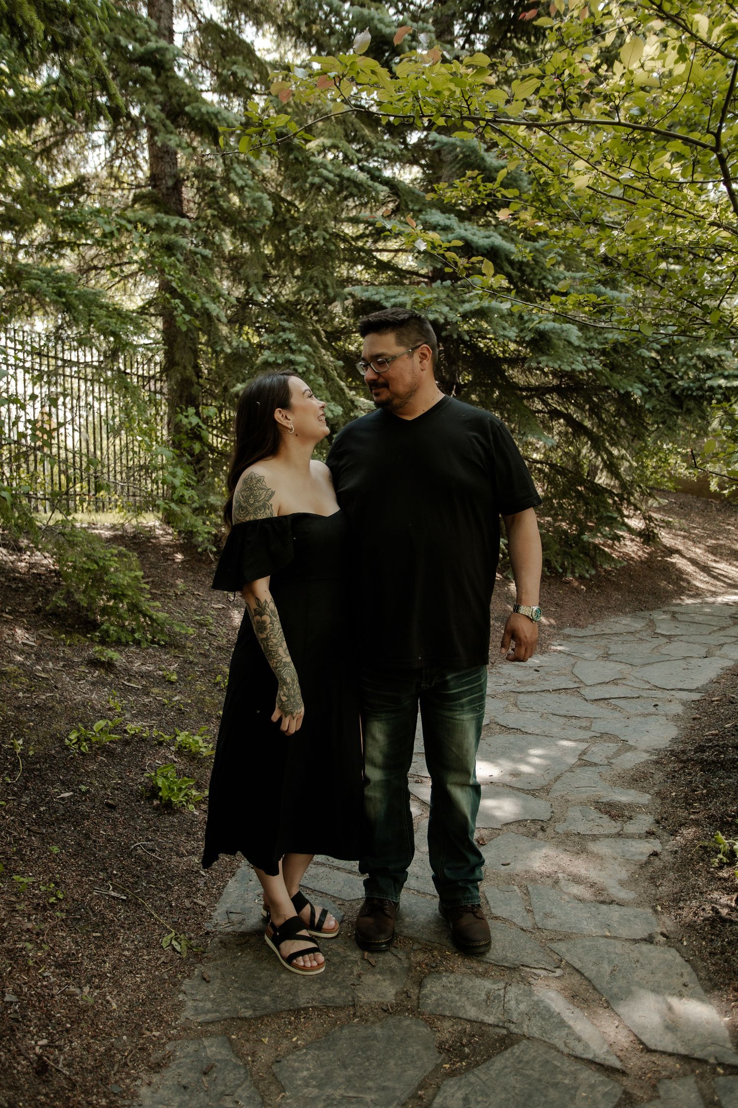 A couple in matching black outfits share a tender moment on a stone path surrounded by lush green trees.