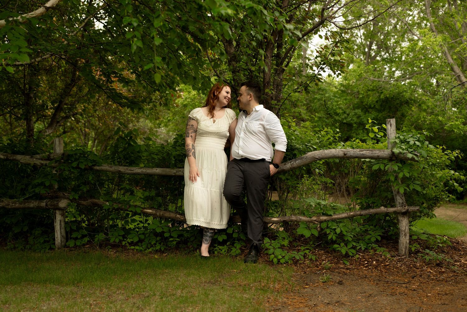 Couple in wedding attire leaning against rustic wooden fence surrounded by lush green trees in outdoor garden setting.