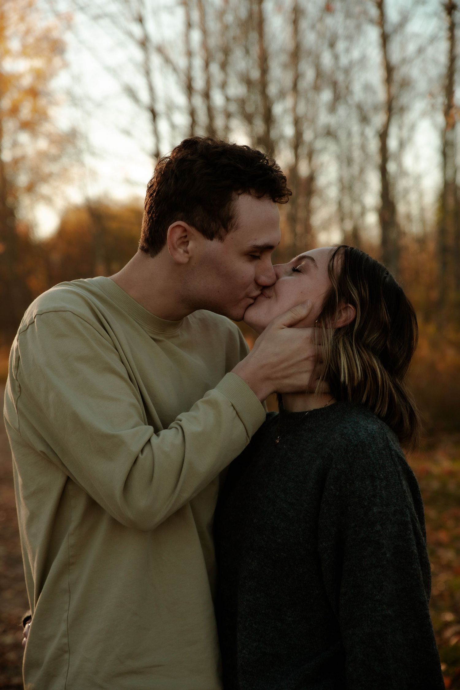 Couple sharing a tender kiss outdoors in a fall forest during golden hour.