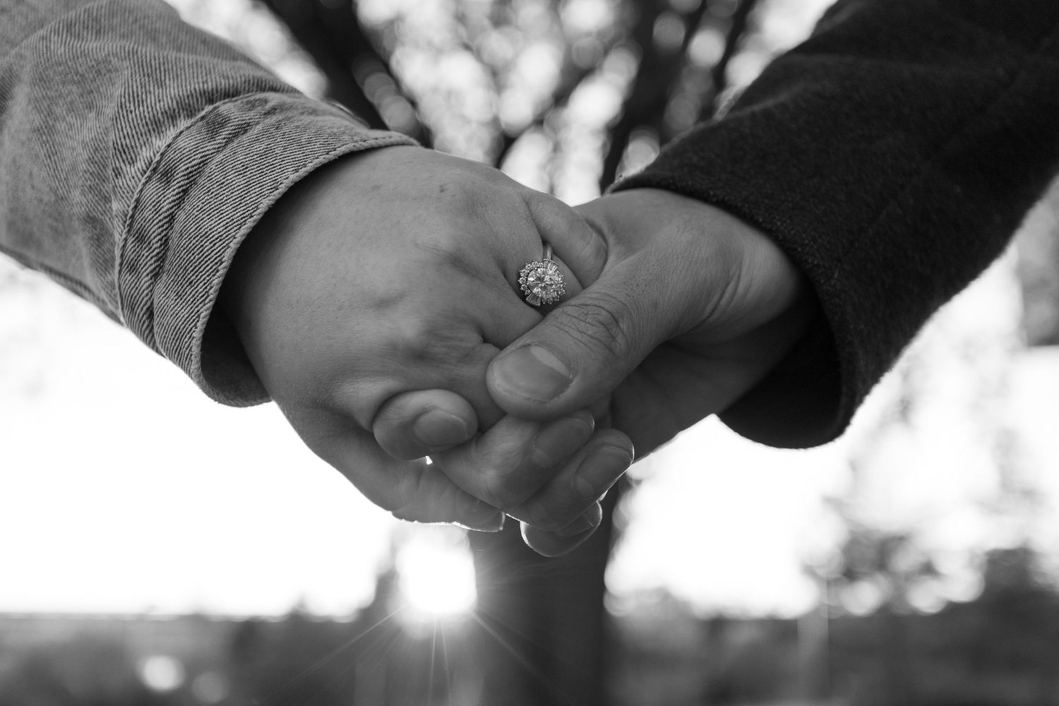 Black and white photo of two people holding hands, one wearing a ring, with blurred trees in the background.