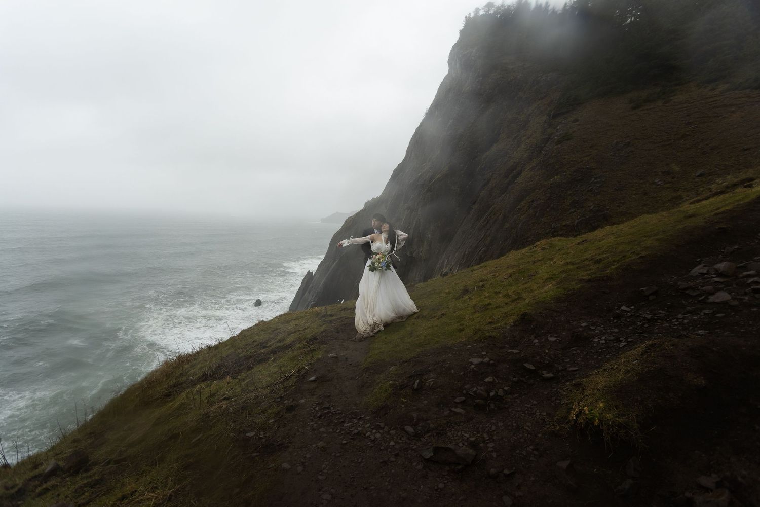 A bride in a white gown stands on a misty coastal cliff with dramatic ocean waves and rocky cliffs in the background.