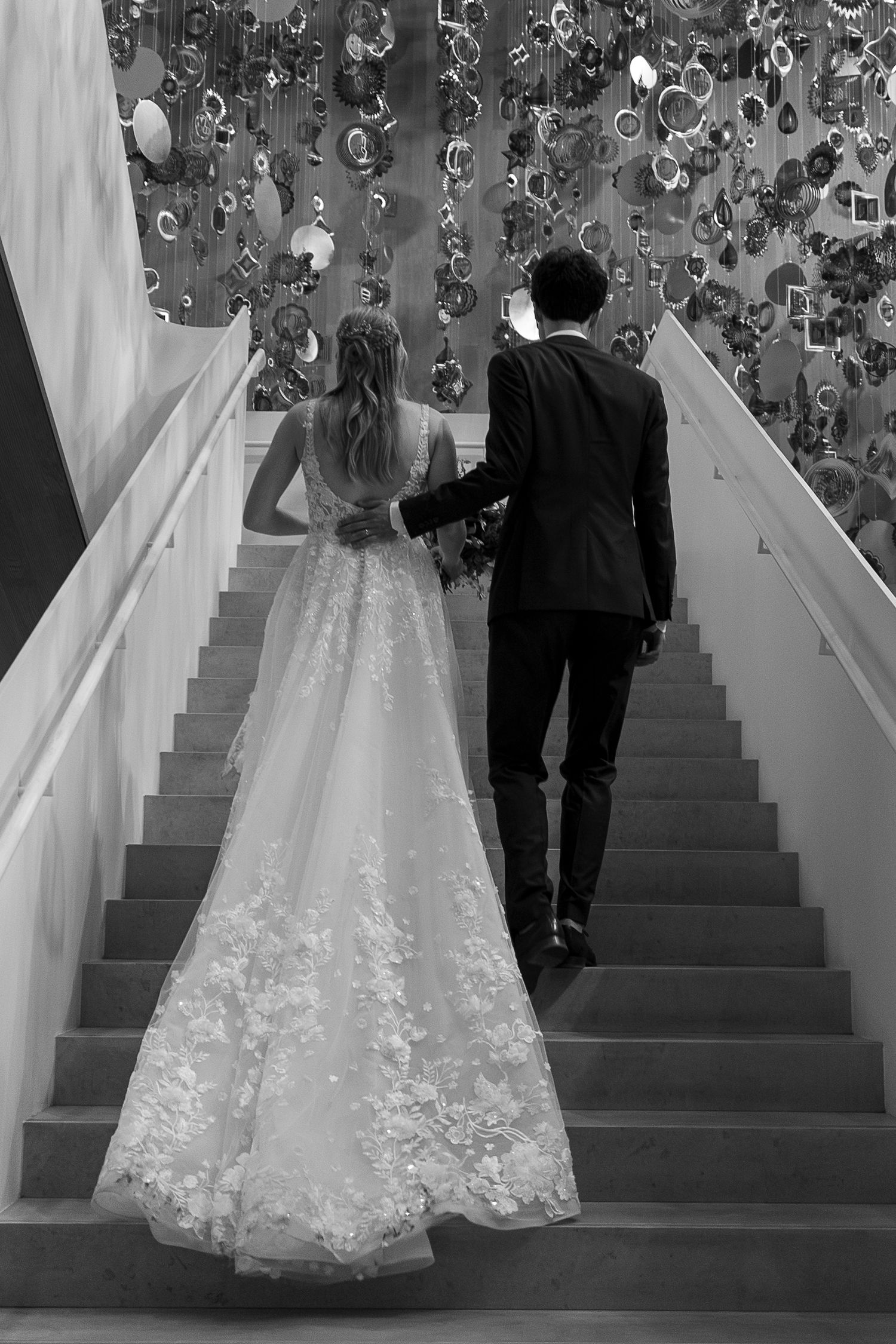 Black and white photo of bride and groom walking up stairs, wedding dress with lace train trailing behind.