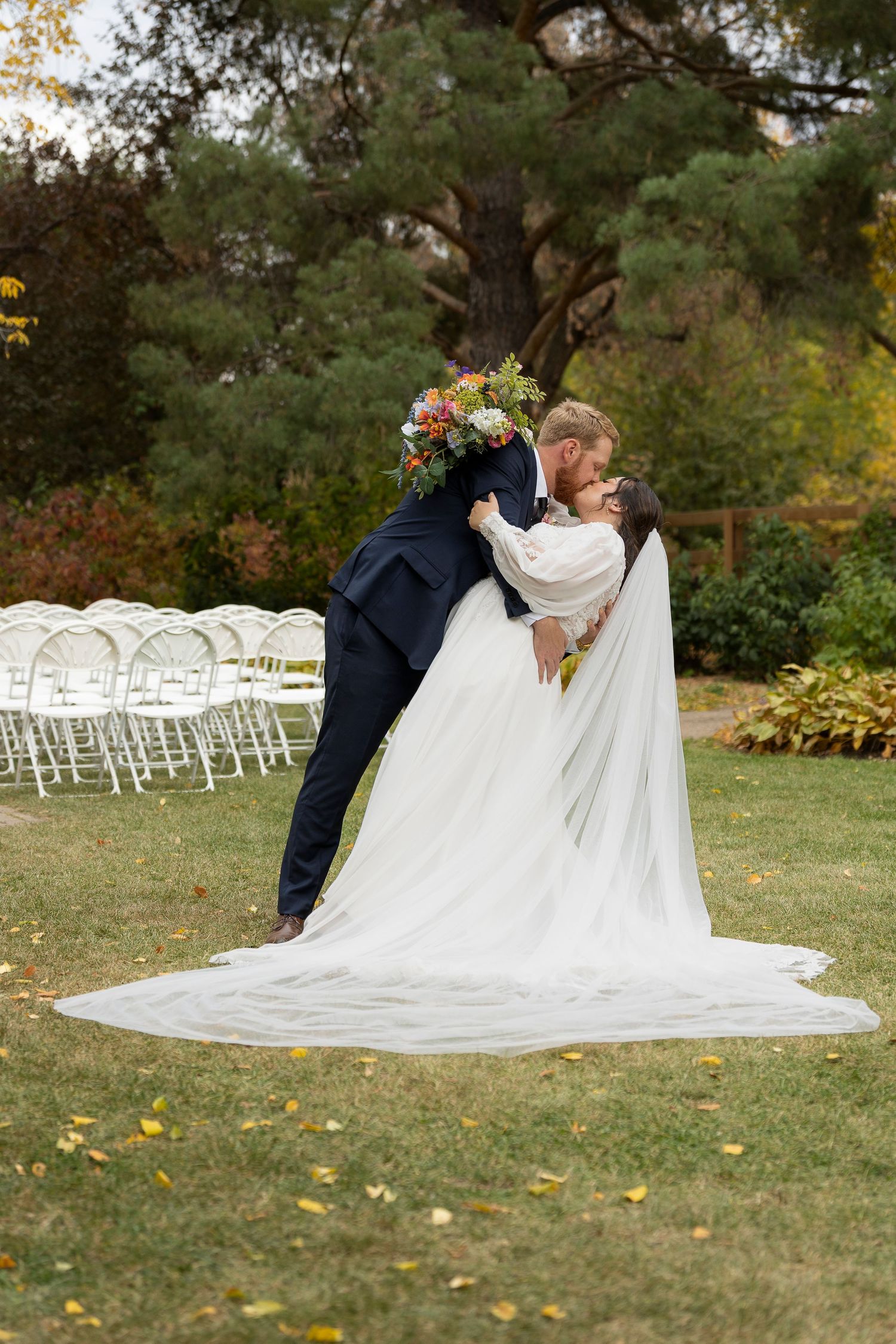 Groom dips bride for a kiss outdoors at garden wedding ceremony, bride in white gown with long veil holding colorful bouquet.